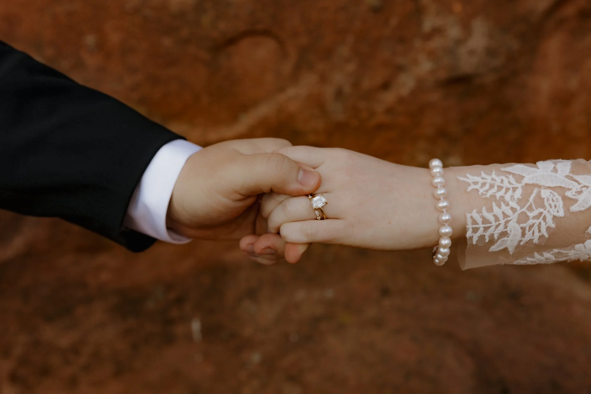A close-up of a couple holding hands during a wedding ceremony, with the bride wearing a lace dress and a pearl bracelet, and the groom in a black suit, against a brownish background.