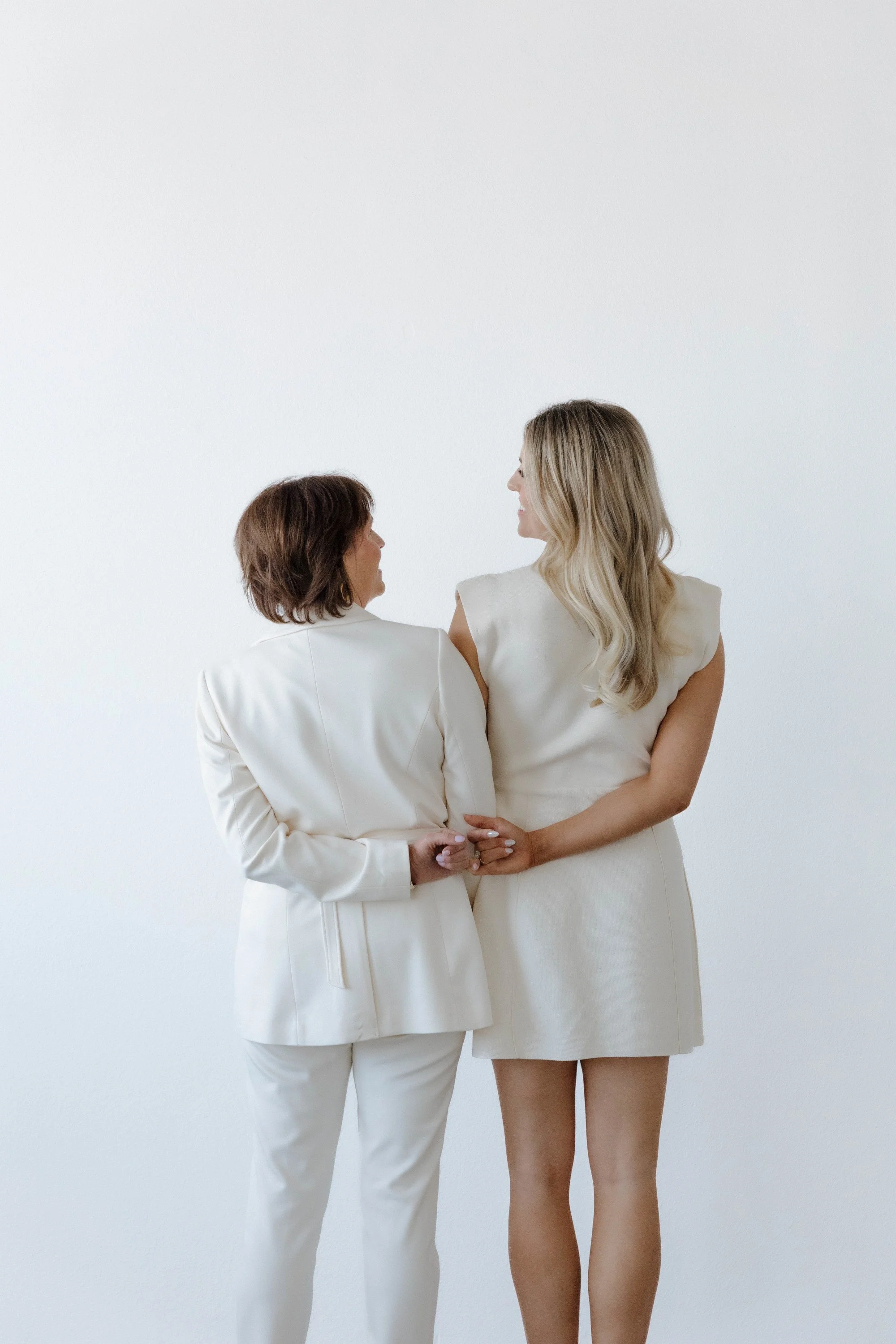 Two women standing together, holding hands, and smiling at each other, dressed in white outfits against a plain white background.