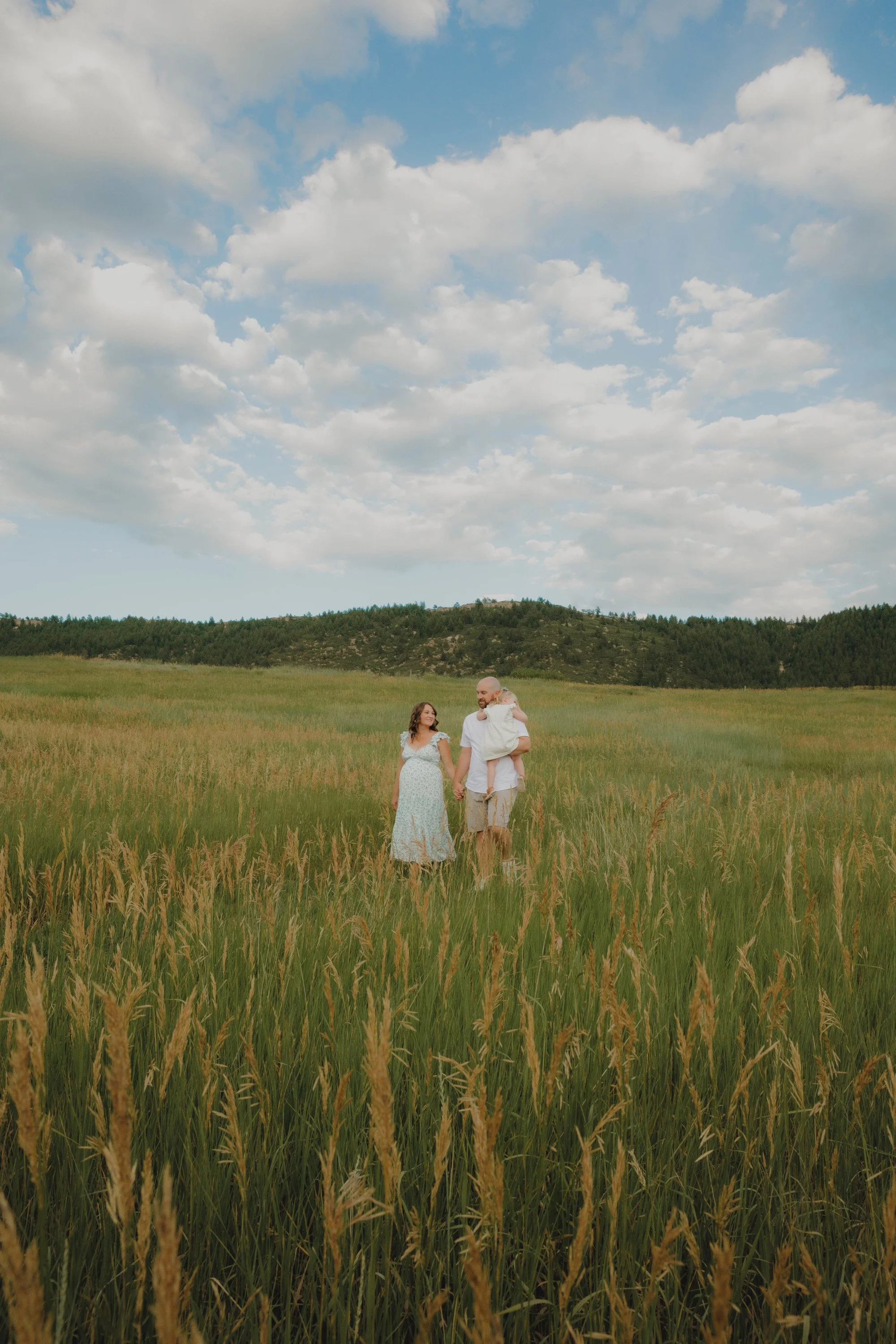 A family of three walking through a lush green field with mountains and blue sky with clouds in the background.