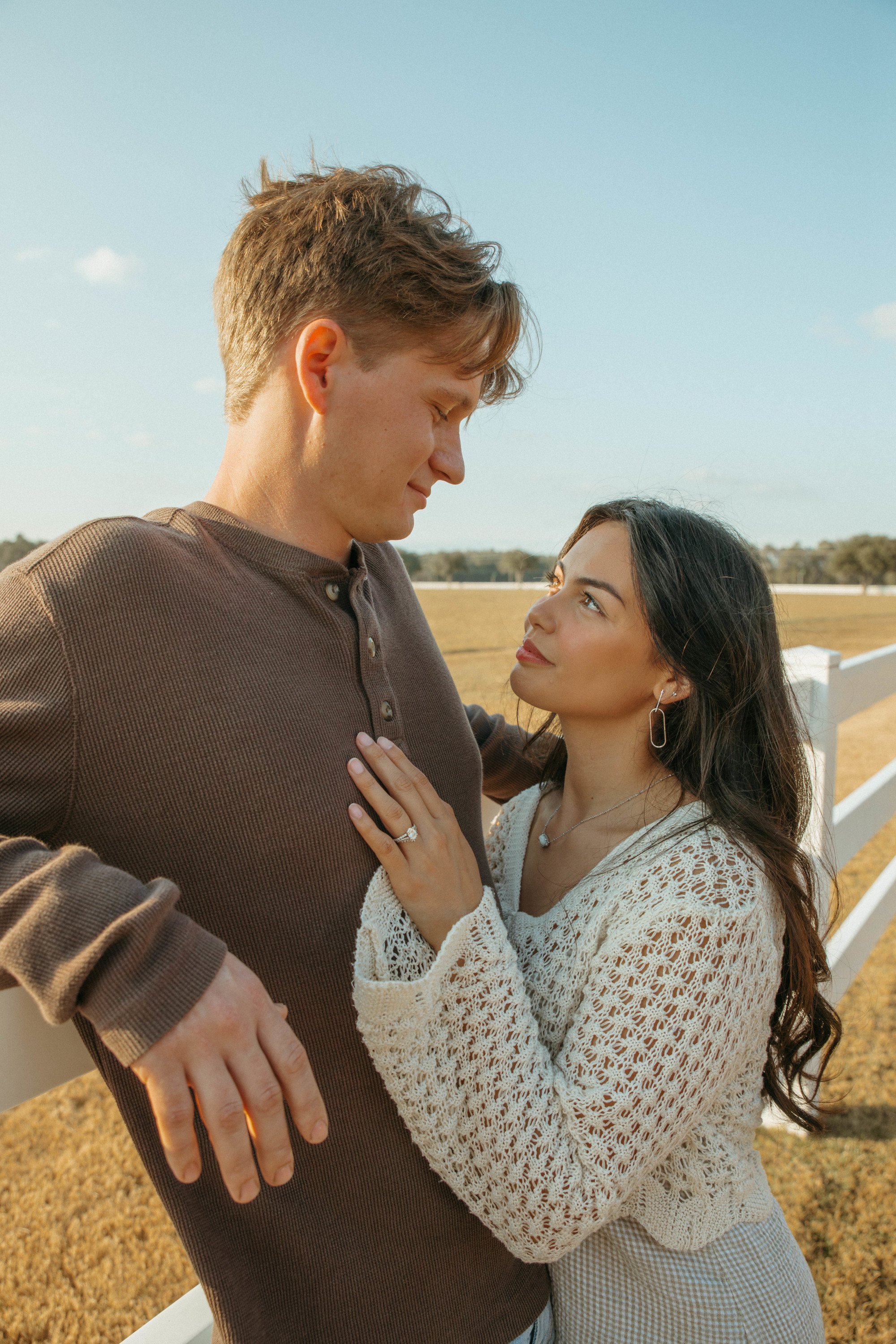 A couple standing outdoors in a field, looking at each other affectionately. The man is wearing a brown long-sleeve shirt and has light brown hair. The woman has long dark hair, earrings, and is wearing a white crochet sweater. The woman is touching 