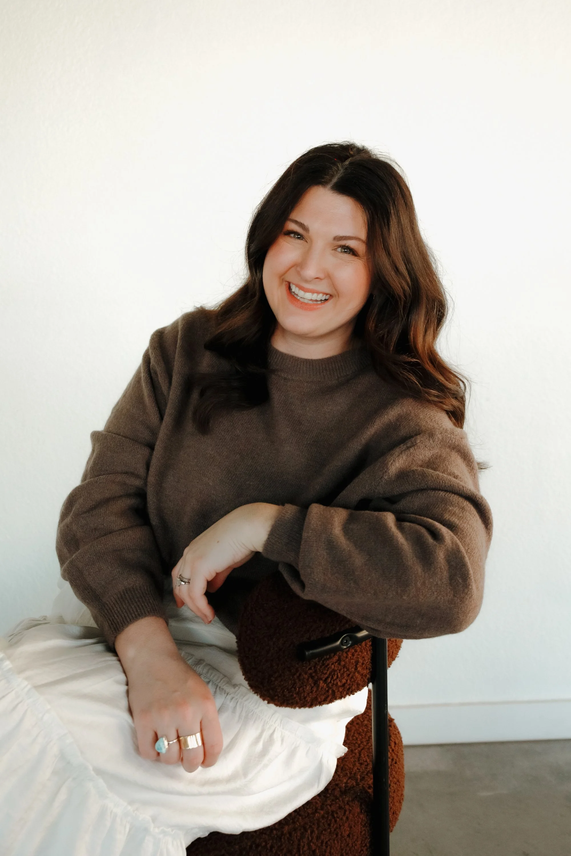 A woman with long dark hair smiling, sitting beside a person with an arm resting on her lap, wearing a brown sweatshirt.