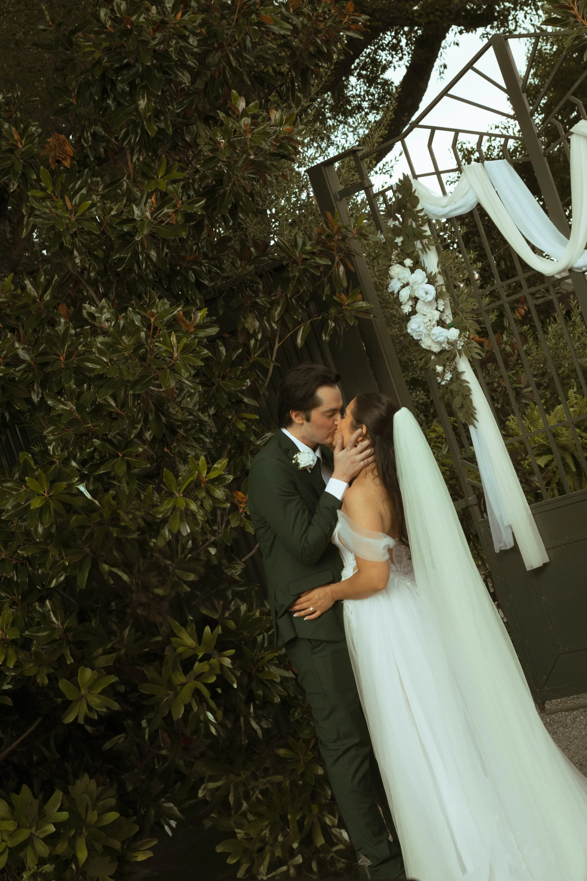 A bride and groom sharing a kiss outdoors at their wedding, with lush greenery and floral decoration behind them.