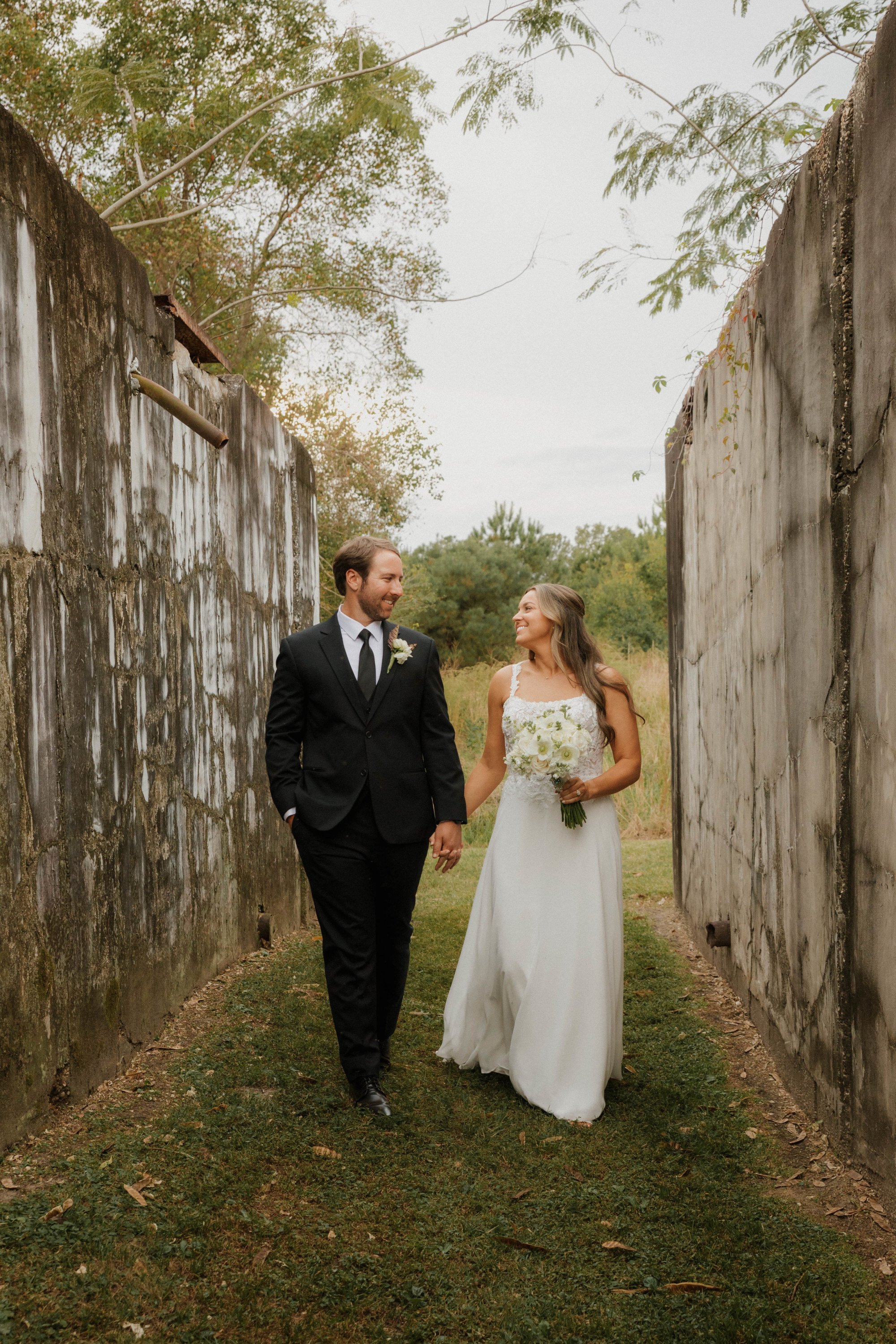 A newlywed couple holding hands while walking through a narrow outdoor passage between old concrete walls, with greenery and trees in the background on an overcast day.