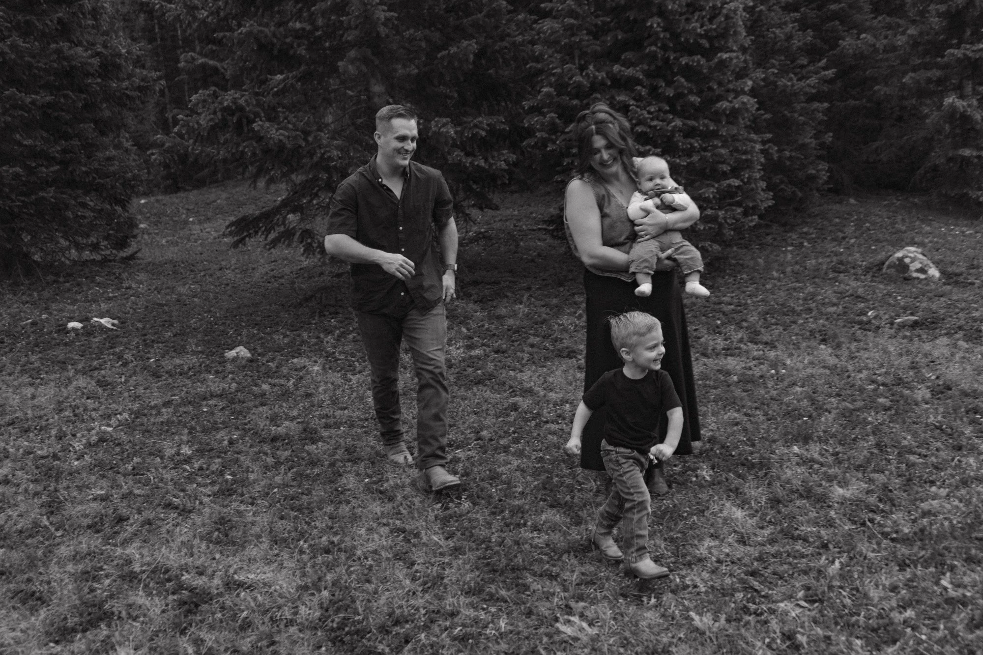 A black and white photo of four people walking outdoors on grass near trees. There is a smiling woman holding a baby, a smiling young boy, and a man walking beside them.
