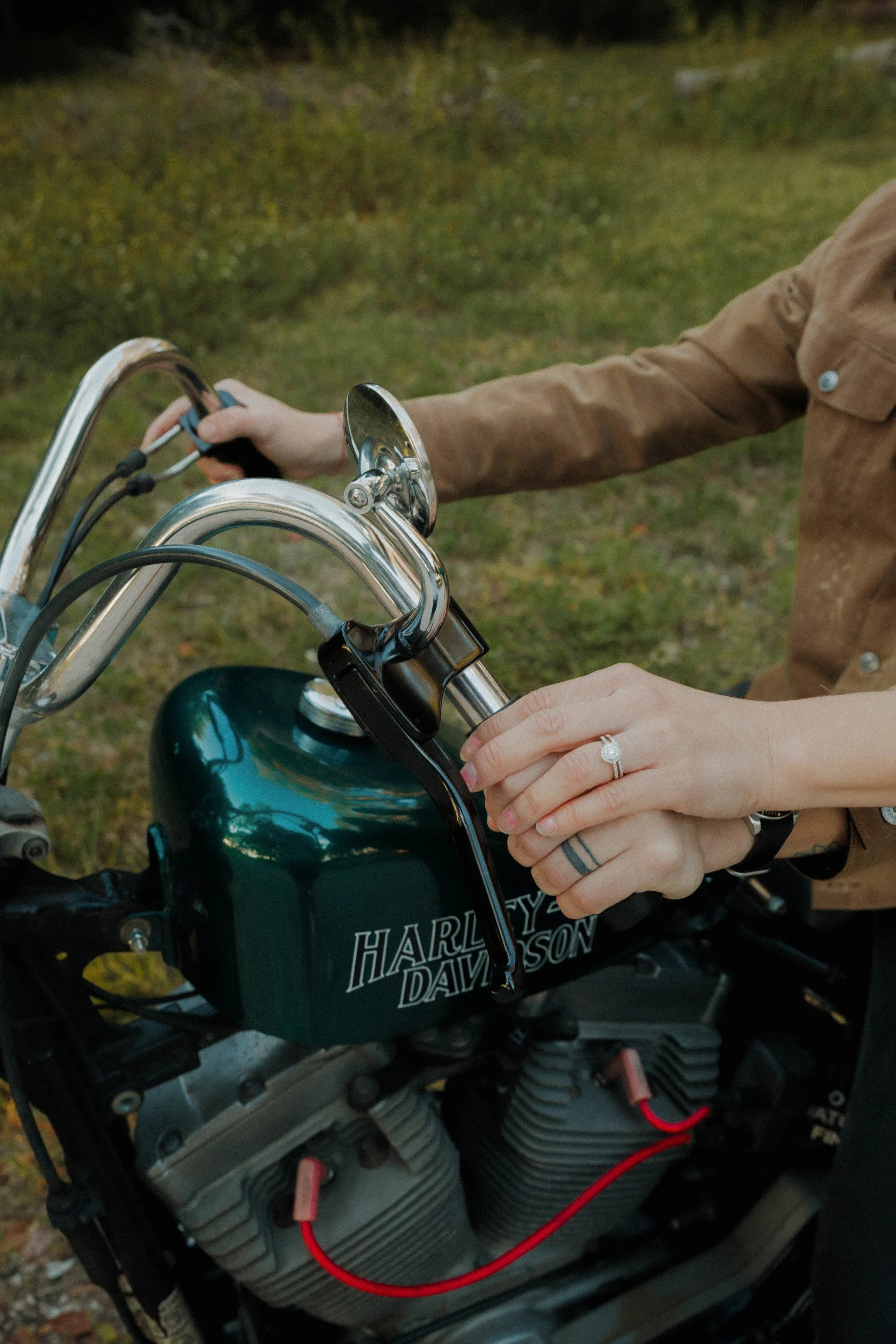 Person wearing a beige jacket with a Harley-Davidson logo on it, starting a motorcycle with an engine and red cables visible, in an outdoor setting with grass and trees in the background.