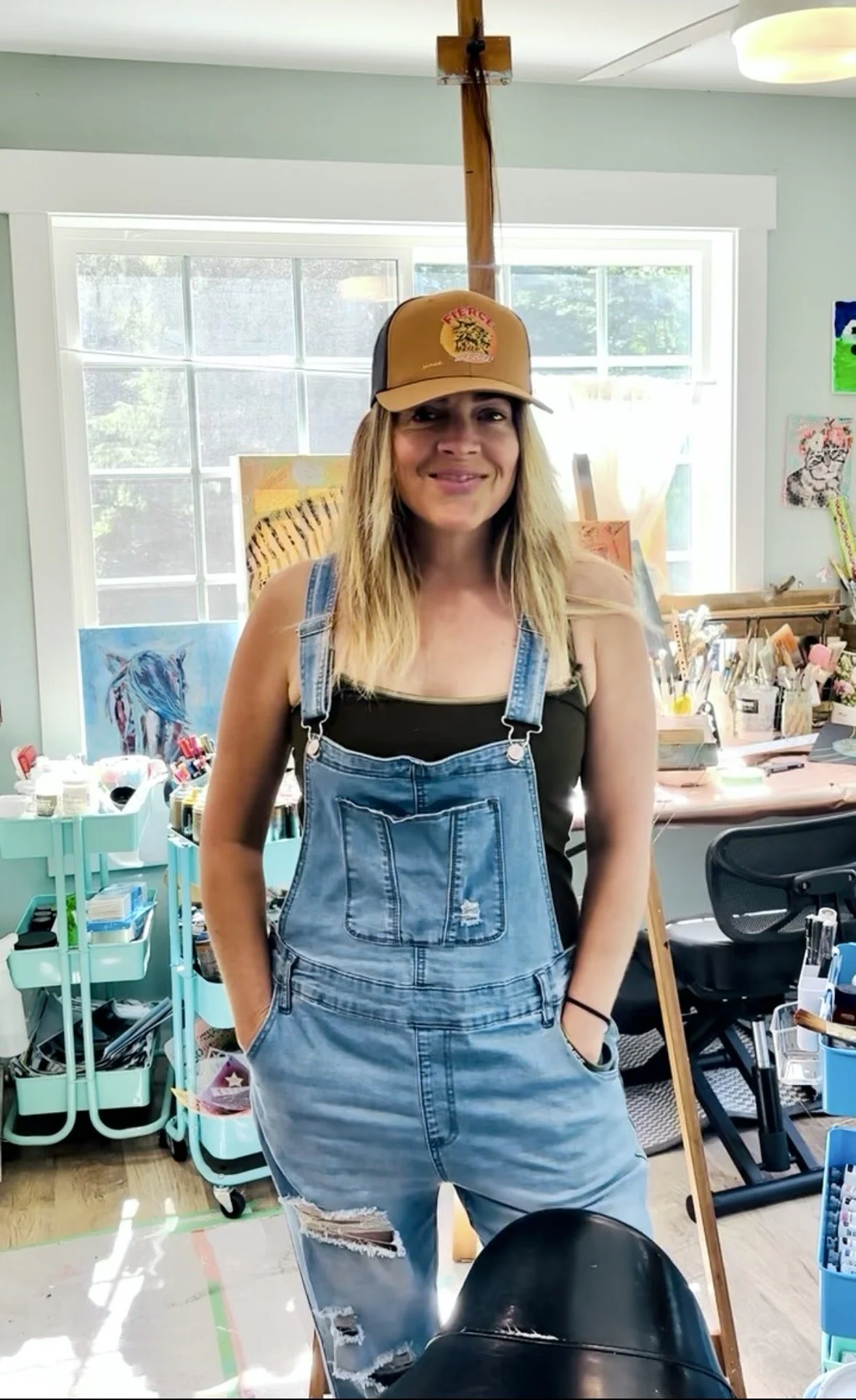 A woman standing in an art studio, wearing a tan trucker hat with a Fierce logo, a black tank top, and distressed denim overalls. The studio has colorful artwork on the walls, art supplies on tables, and a large window in the background.