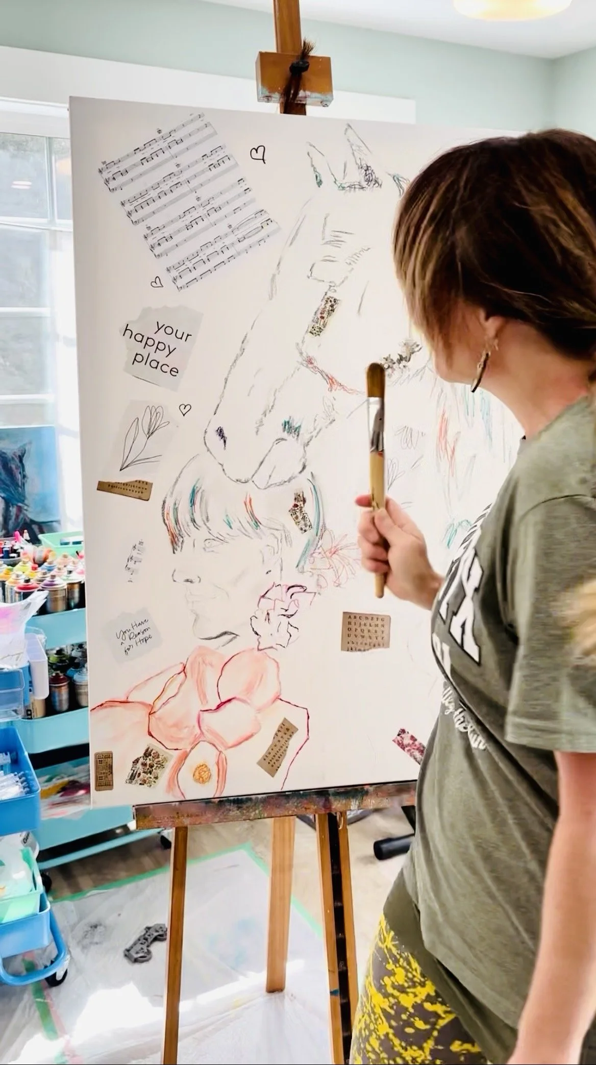 Sally Kaye the Artist who paints expressive and emotional works from her love of life and nature holding a paintbrush in front of a canvas with sketches of animal faces, musical notes, and cut-out words and phrases in an art studio.
