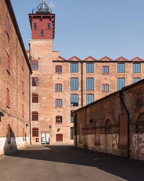 View of old brick building with tower and windows, along with surrounding brick structures under a clear blue sky.