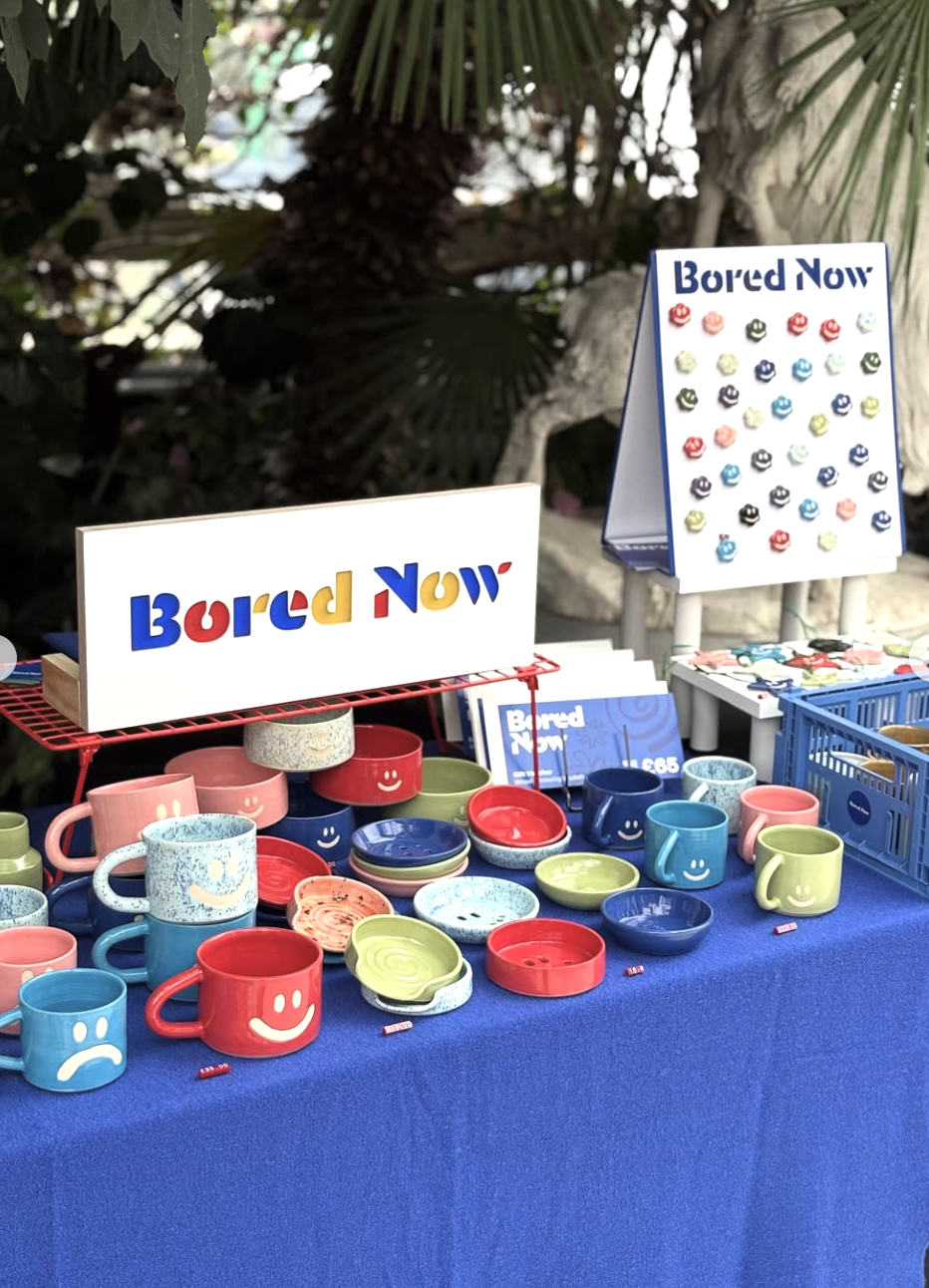 Display table with colorful mugs and bowls, some with smiley faces, and signs that read 'Bored Now'; the sign on the table has the same phrase, but printed upside down.