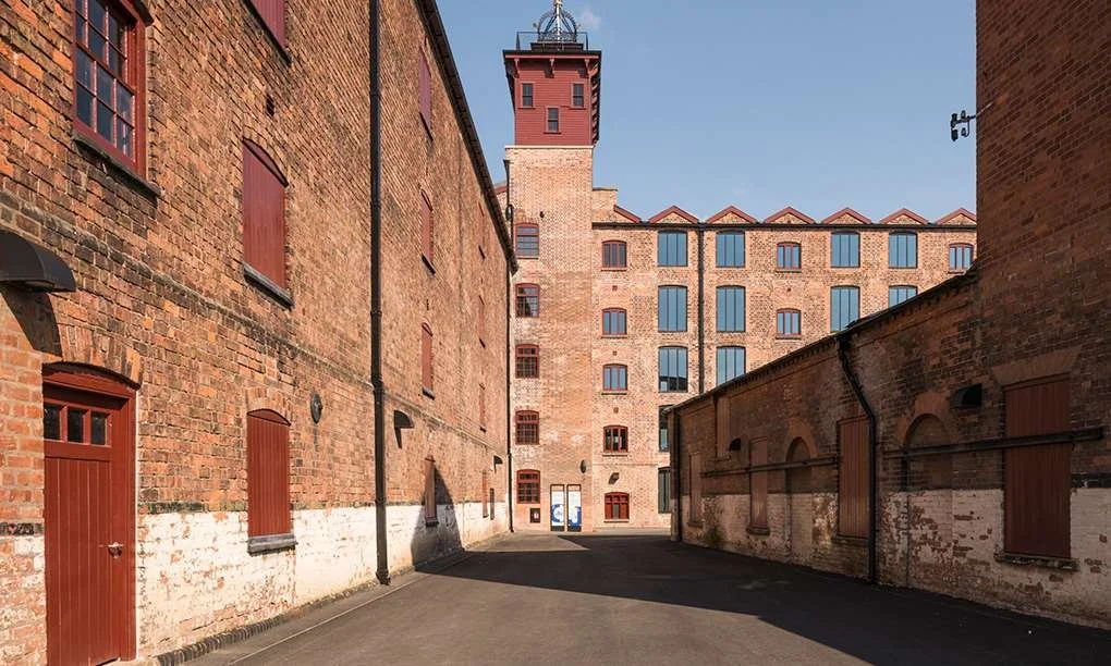 View of a narrow alleyway between brick buildings with a taller brick building and a tower in the background under a clear blue sky.
