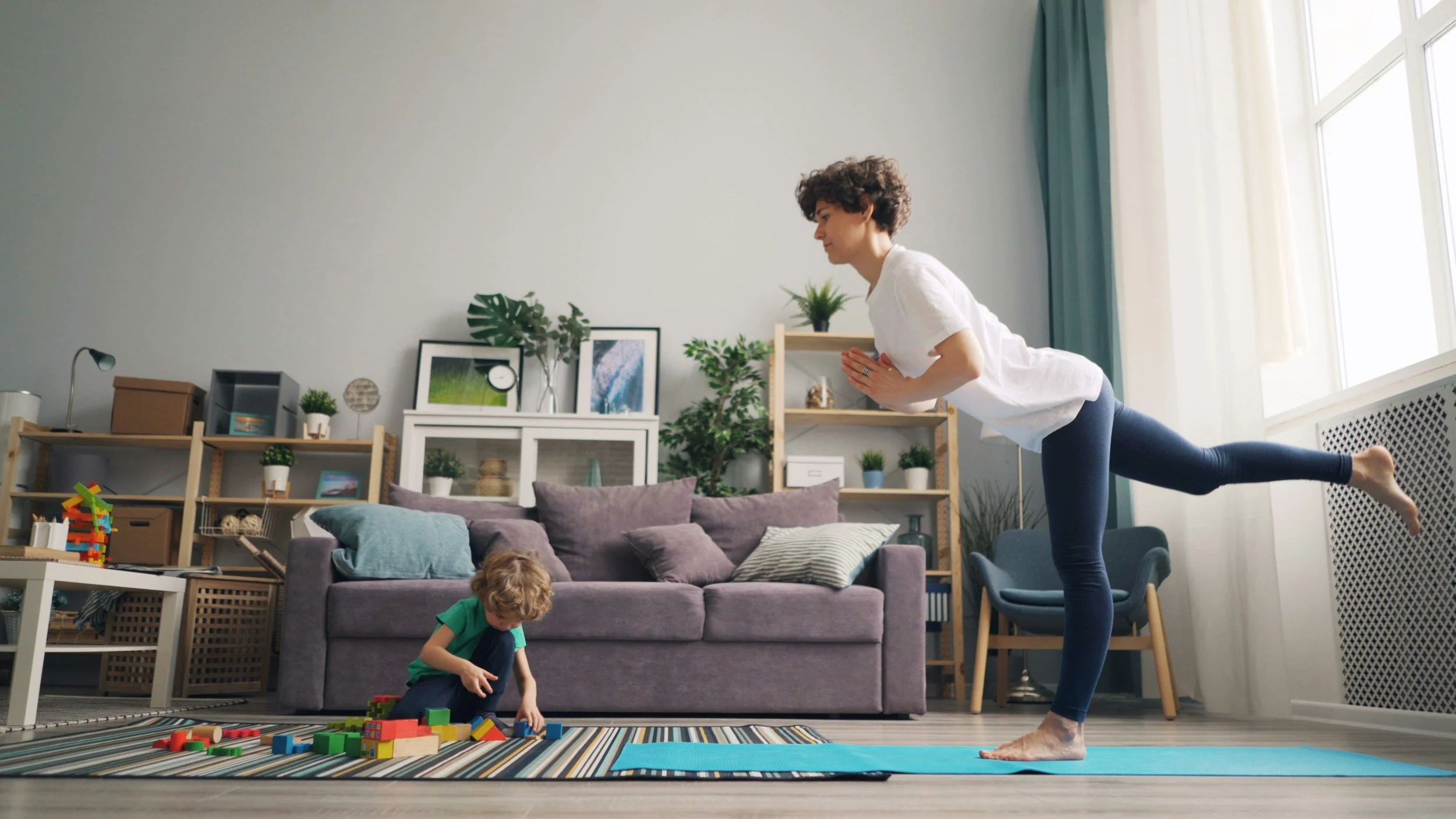 A woman doing yoga on a blue mat in a living room, with two children playing on the colorful rug nearby. The room has a gray couch, multiple shelves with decorative items, plants, and art on the wall. Natural light streams in through a large window with curtains.