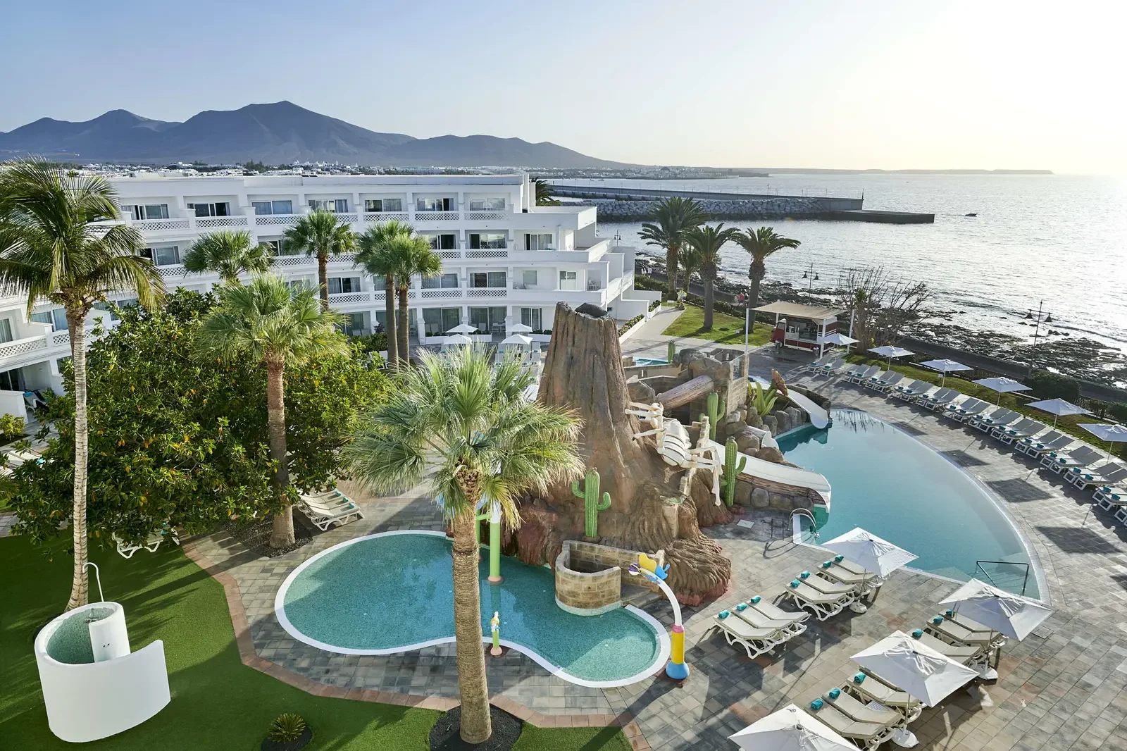 Luxe resortzwembad met een bergachtige achtergrond, palmbomen en uitzicht op de oceaan, met ligbedden en parasols rondom het zwembad.