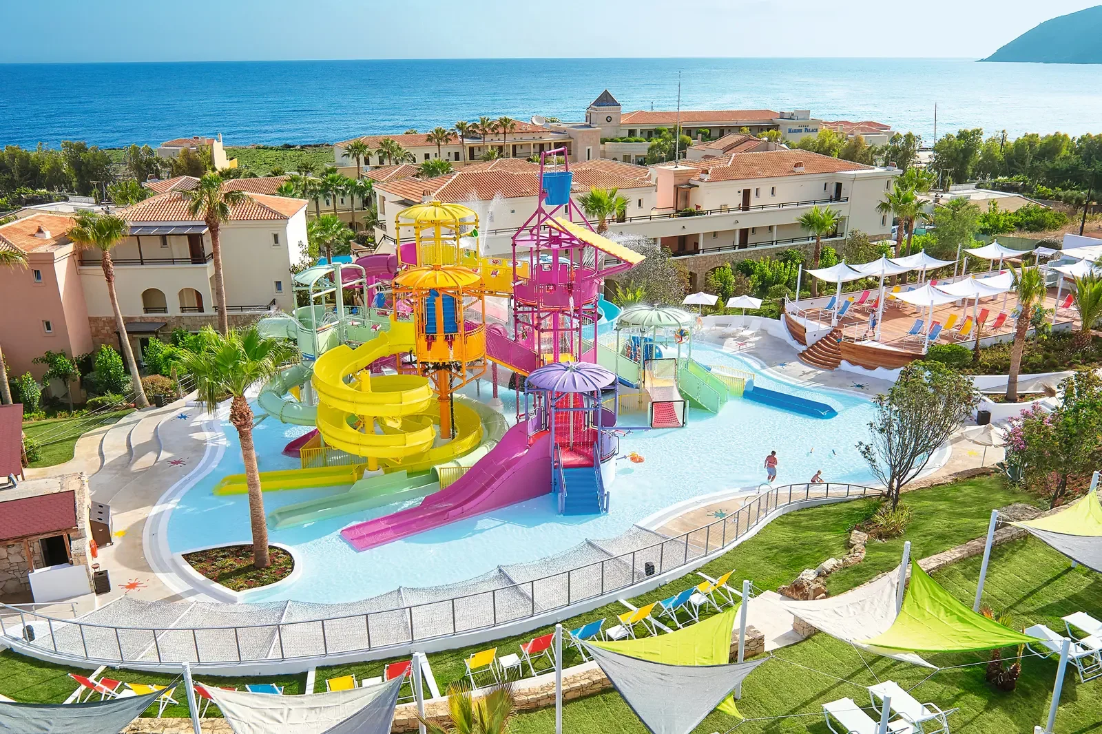 Een kleurrijk waterpark met verschillende glijbanen, ligbedden en zwemmers, gelegen nabij de zee met palmbomen en appartementen eromheen.