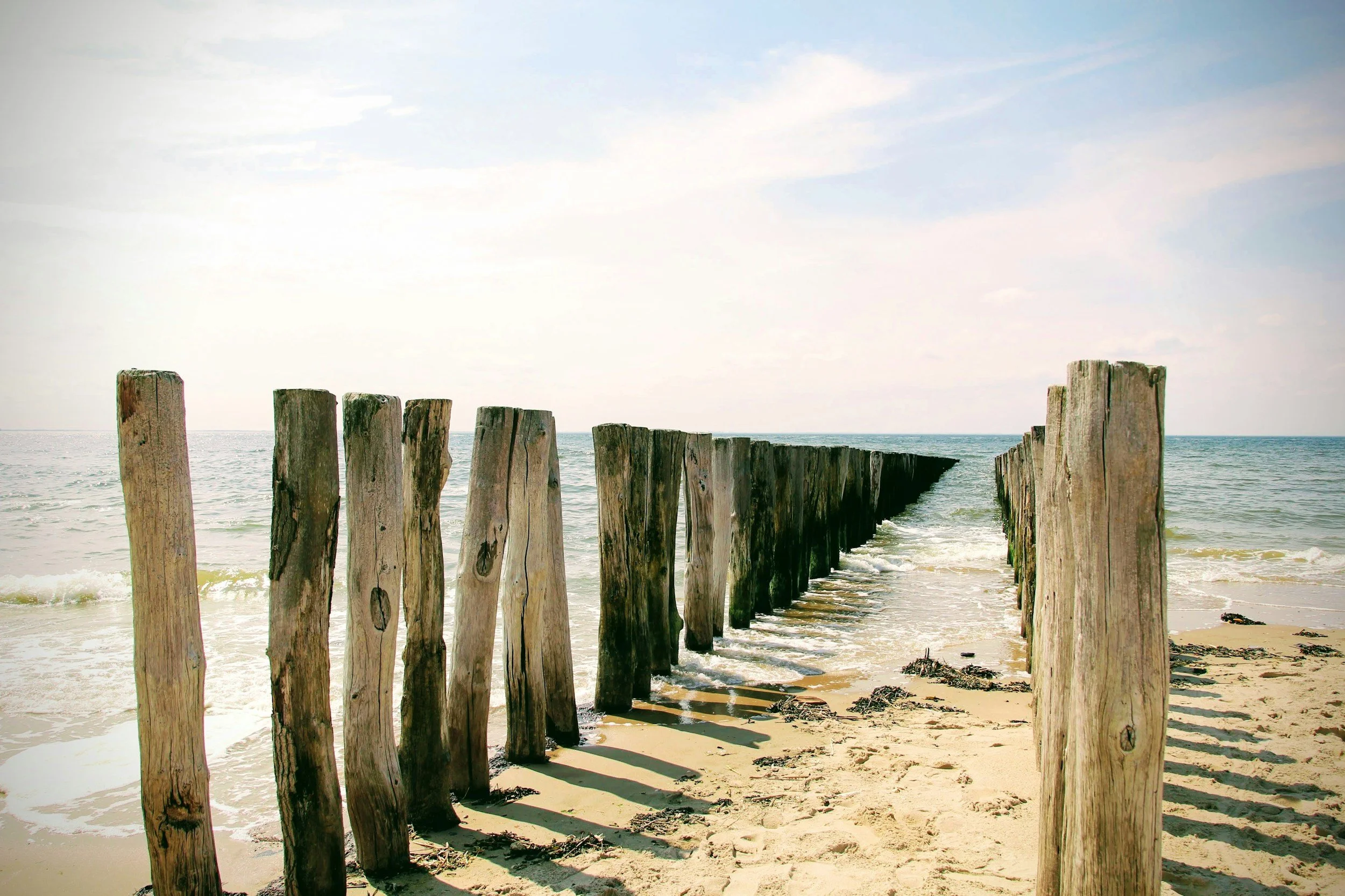 Een houten pier die in de oceaan ligt, met gerotte houten palen die door het water steken, onder een gedeeltelijk bewolkte hemel.