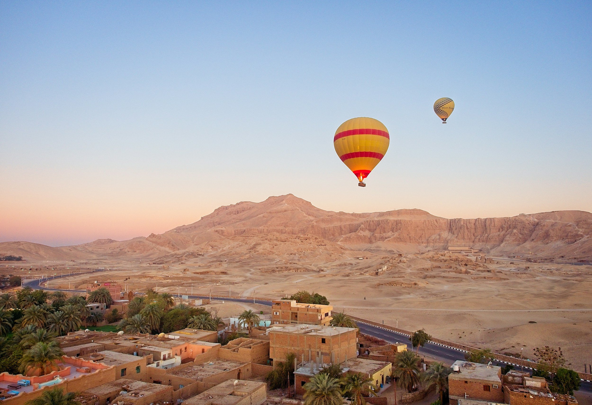 Luchtfoto van een landschap met twee ballonnen in de lucht en een droge, bergachtige omgeving met een klein dorp en palmbomen op de voorgrond.