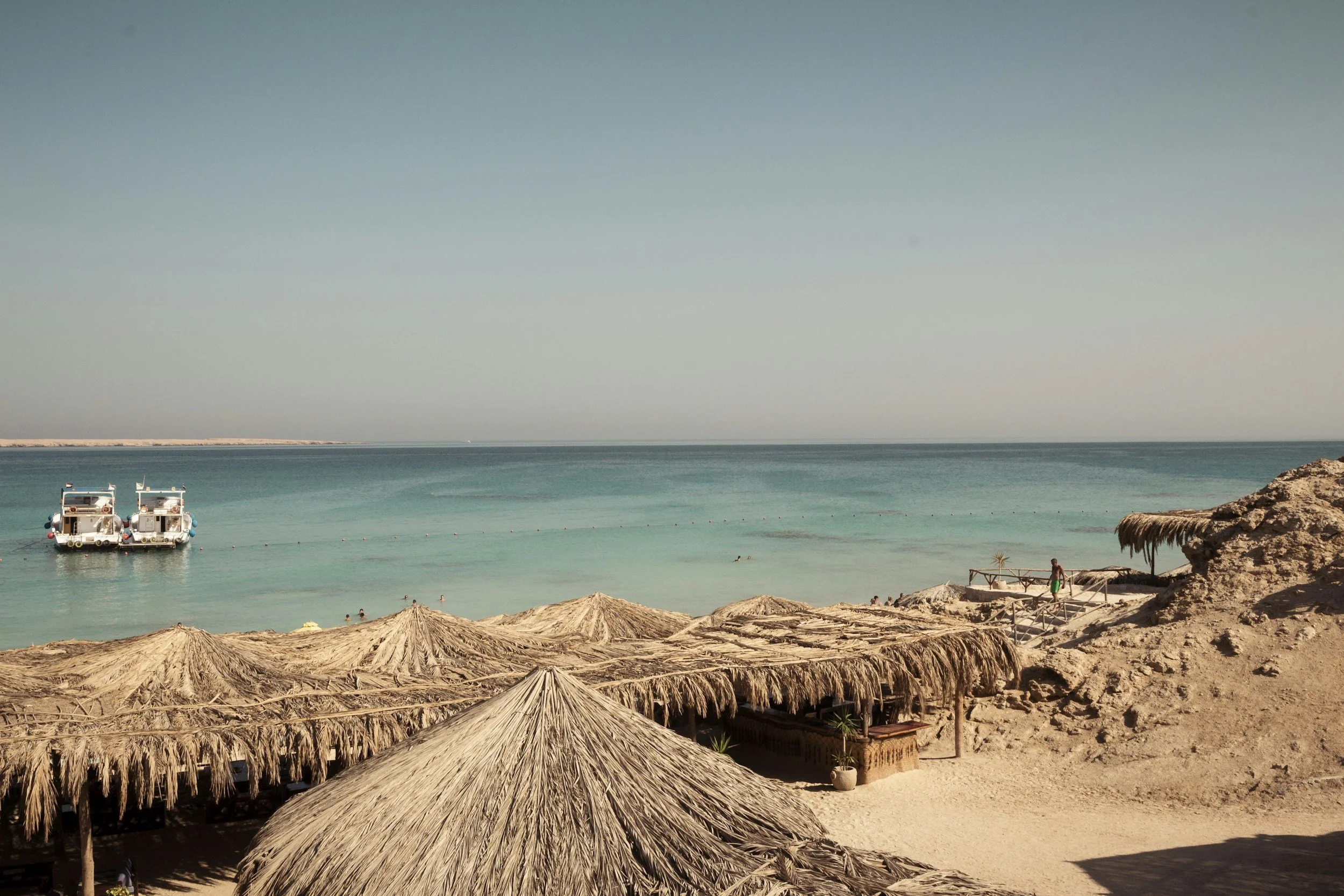 Een strand met rieten parasols, mensen in het water en een blauwe zee onder een heldere hemel.