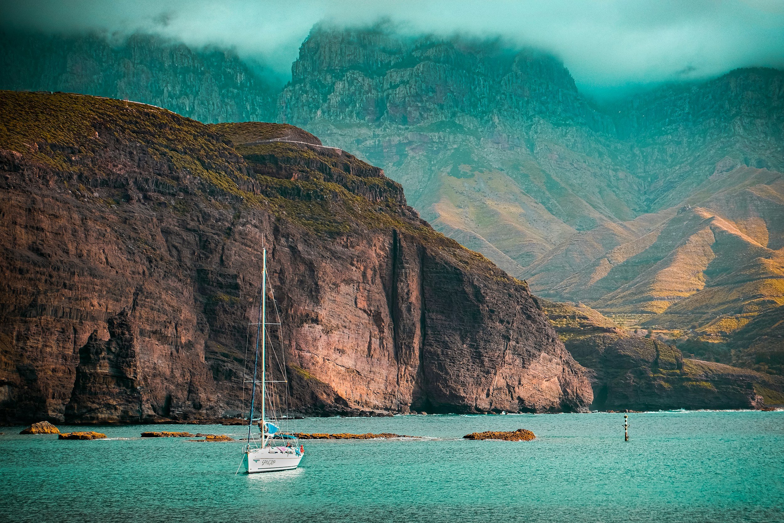 Een zeilboot op turquoise water voor rotsachtige kliffen met groene vegetatie en laaghangende bewolking.