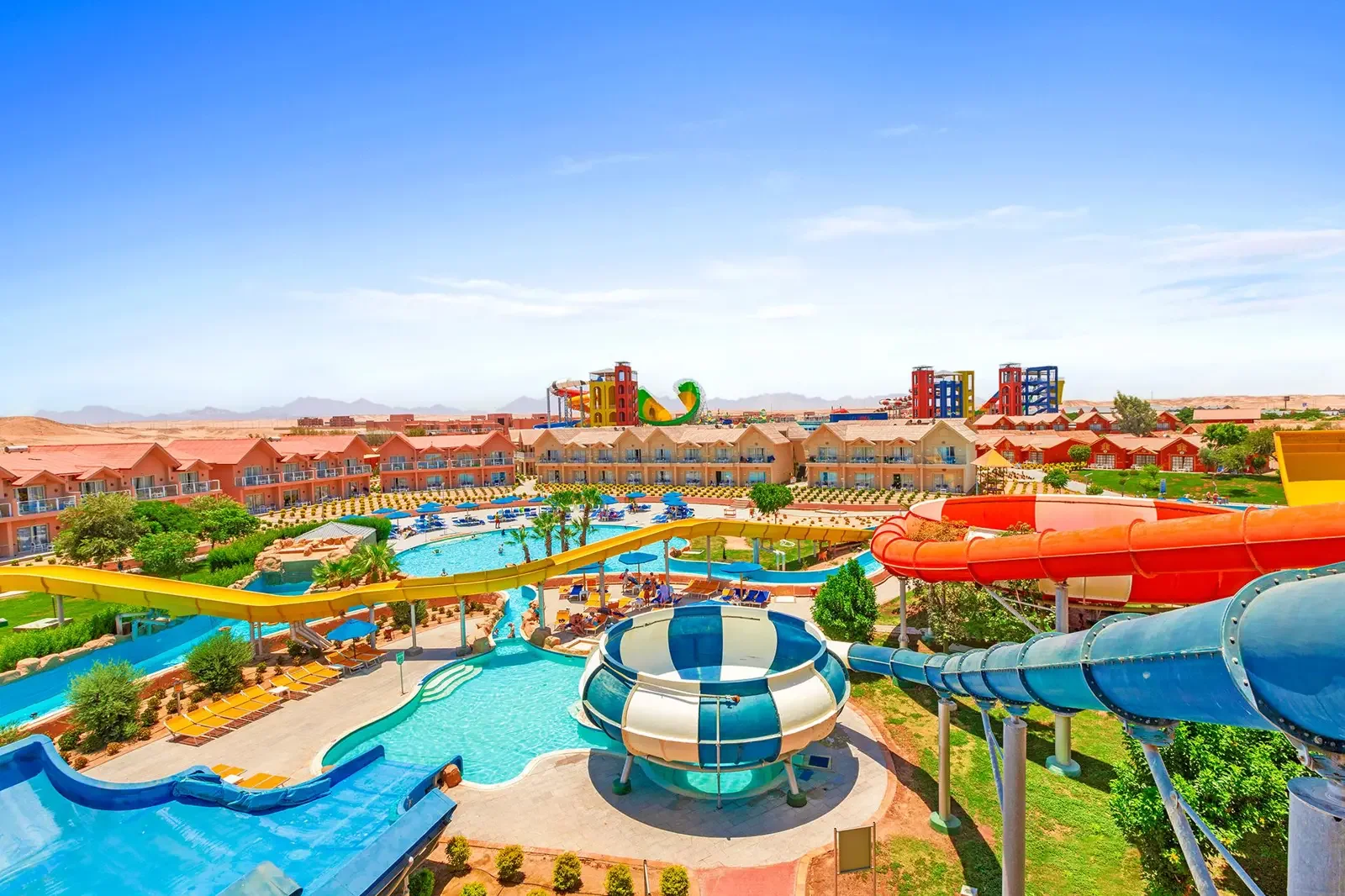 Gezicht op een waterpark met glijbanen, zwembad en omliggende gebouwen onder een blauwe hemel.