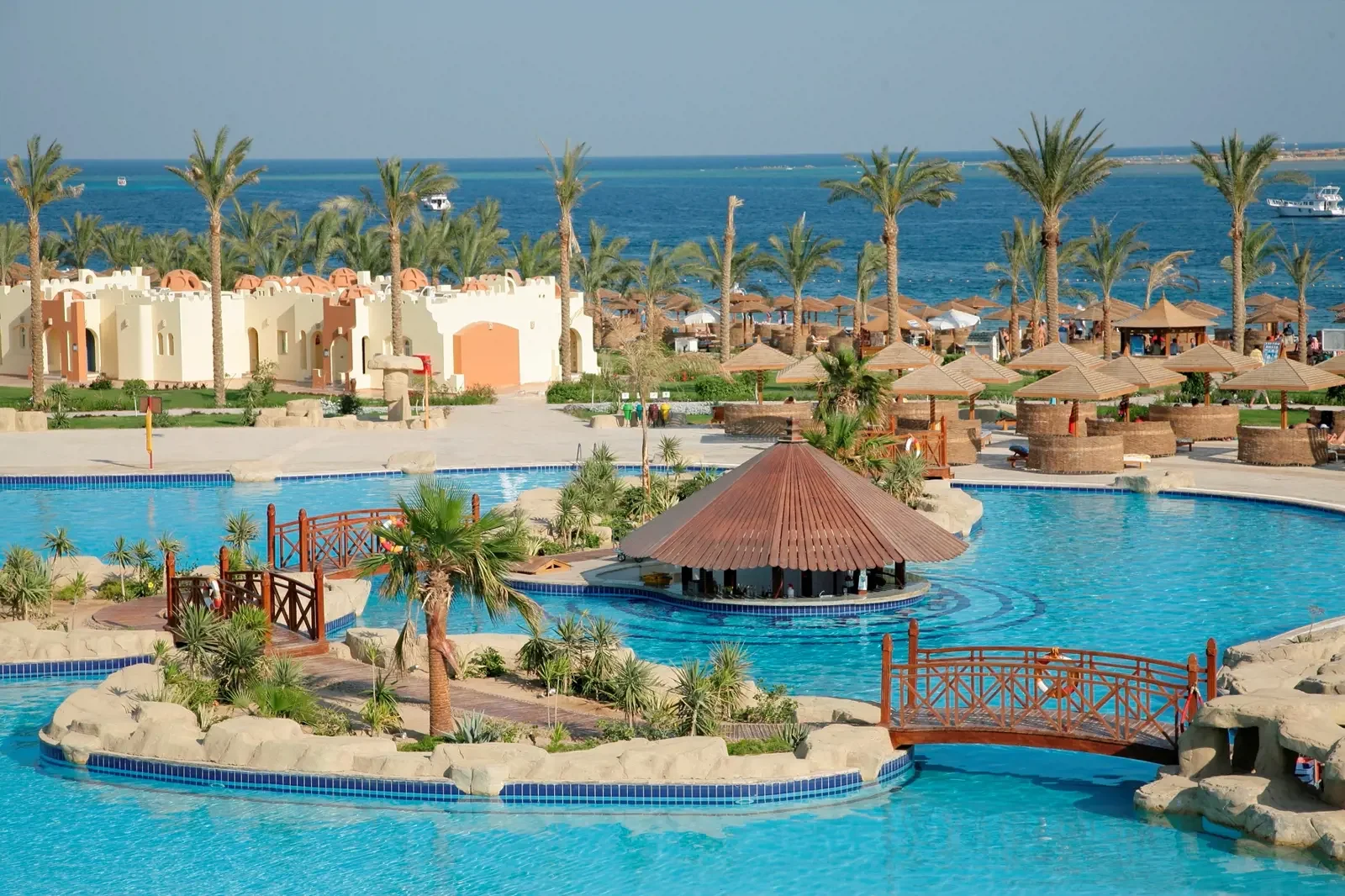 Luxe resort met zwembaden, palmbomen, strandbedden en gazebos, met de zee en boten op de achtergrond.