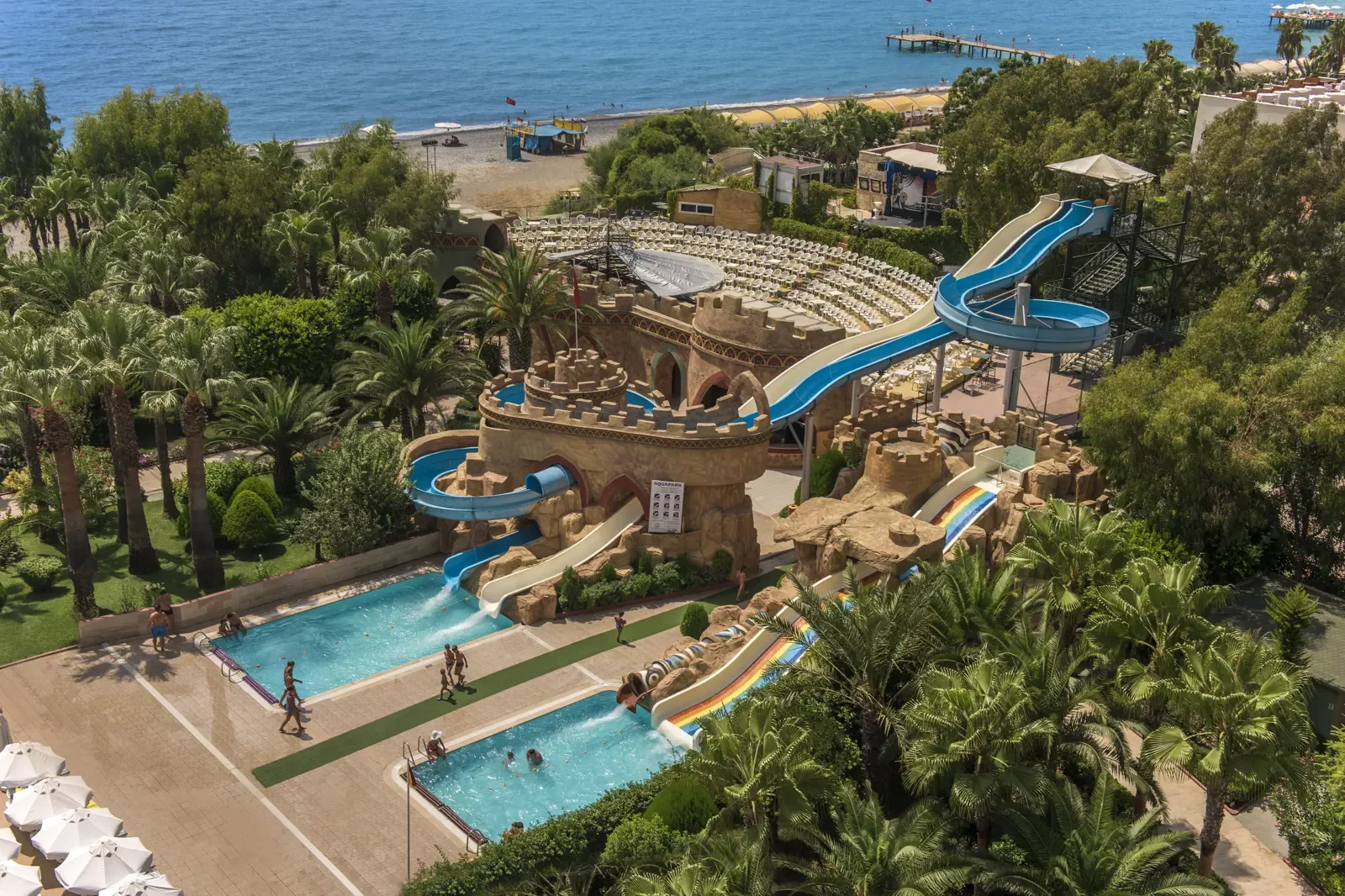 Een waterpark met meerdere glijbanen, een zwembad, omgeven door palmbomen en toeristen, met de zee op de achtergrond.