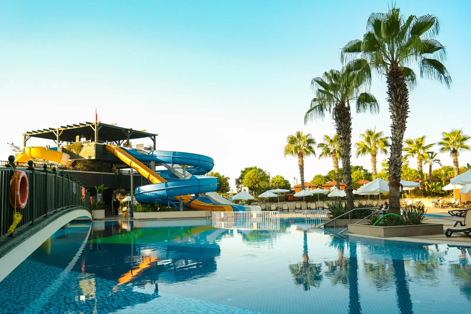 Zwembad met waterglijbanen en ligstoelen onder palm- en andere bomen bij een resort