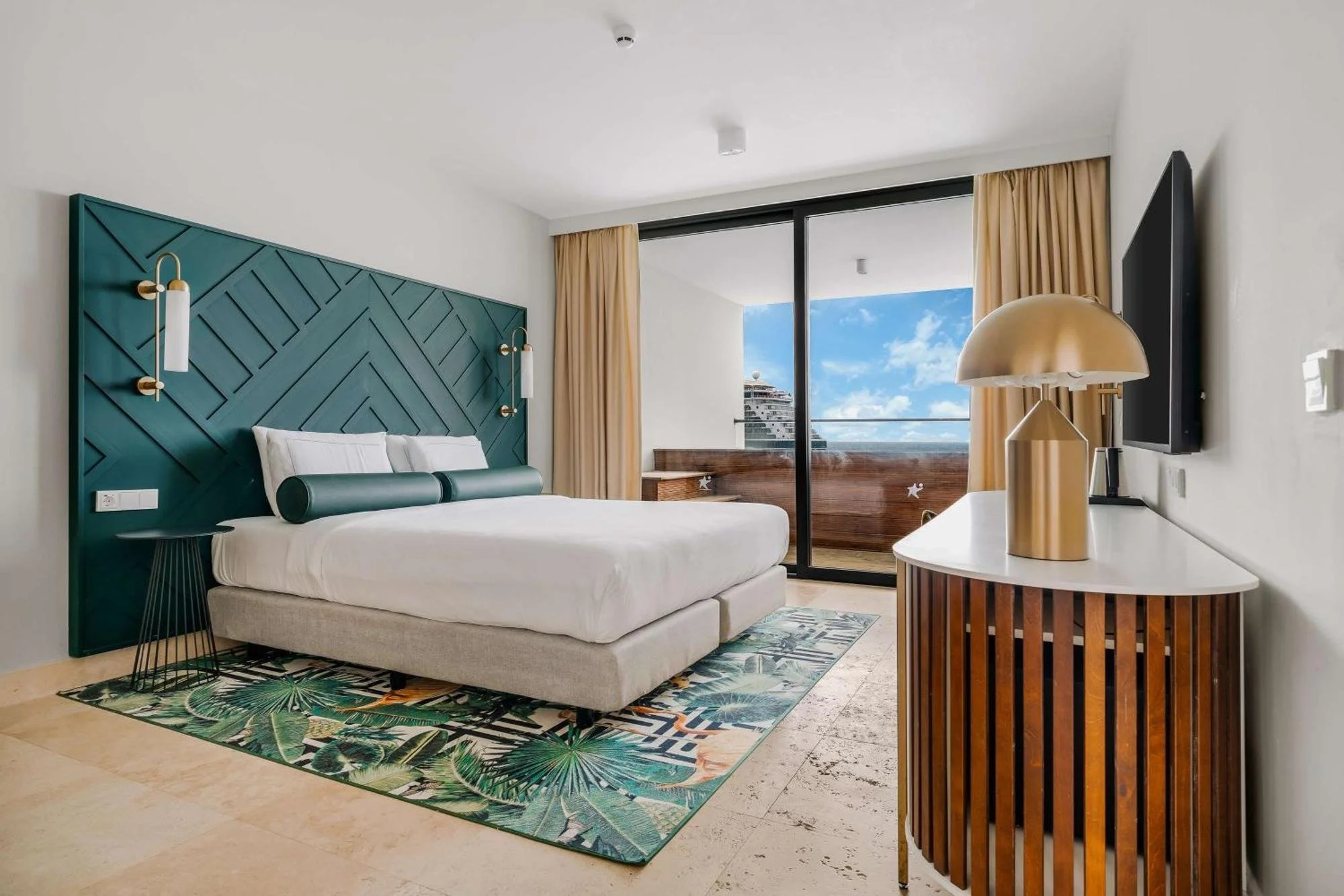Hotelkamer met wit bed, blauwe hoofdtoog met geometrisch patroon, geel gordijn, zwart tv, gouden lamp, balkon met uitzicht op zee en een hotelgebouw.