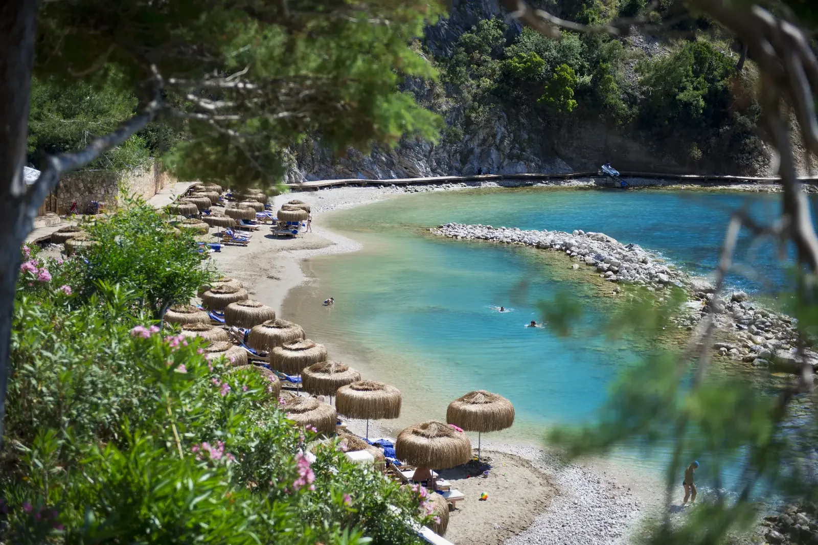 Een klein strand met ligstoelen en parasols, omgeven door groene planten en uitzicht op de zee met enkele mensen die zwemmen, in een rustige en natuurlijke omgeving.