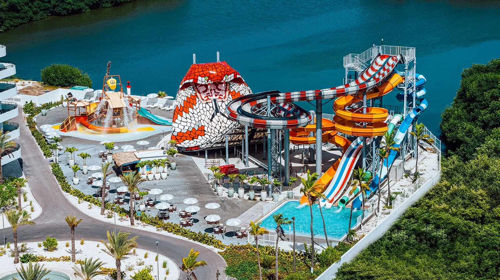 Luchtfoto van een waterpark met meerdere glijbanen die in een grote vijver uitmondt, omringd door bomen en ligstoelen met parasols.