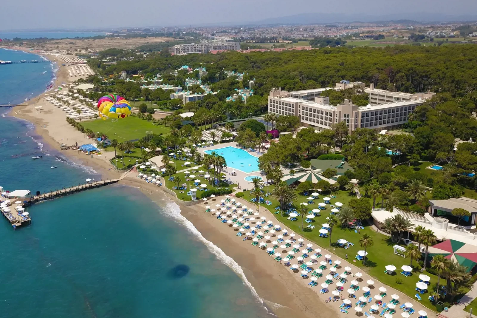 Luchtfoto van een resort met strand, zwembaden, ligbedden en parasols, met groen bebost gebied en een groot gebouw, meerdere hot air ballonnen in de lucht.