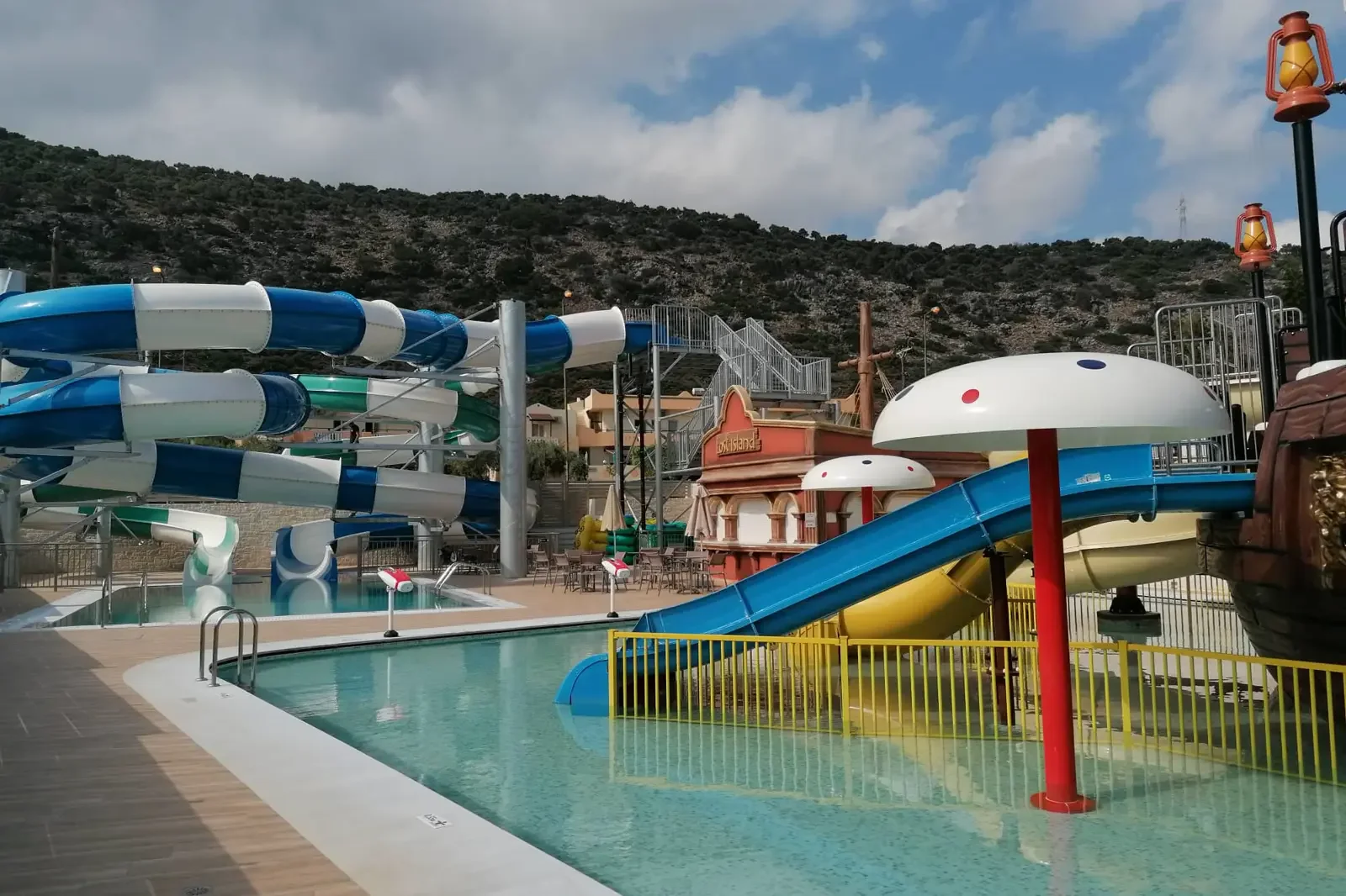 Een waterpark met glijbanen, een mini zwembad en een decor van een schip, met bergen op de achtergrond en bewolkte lucht.