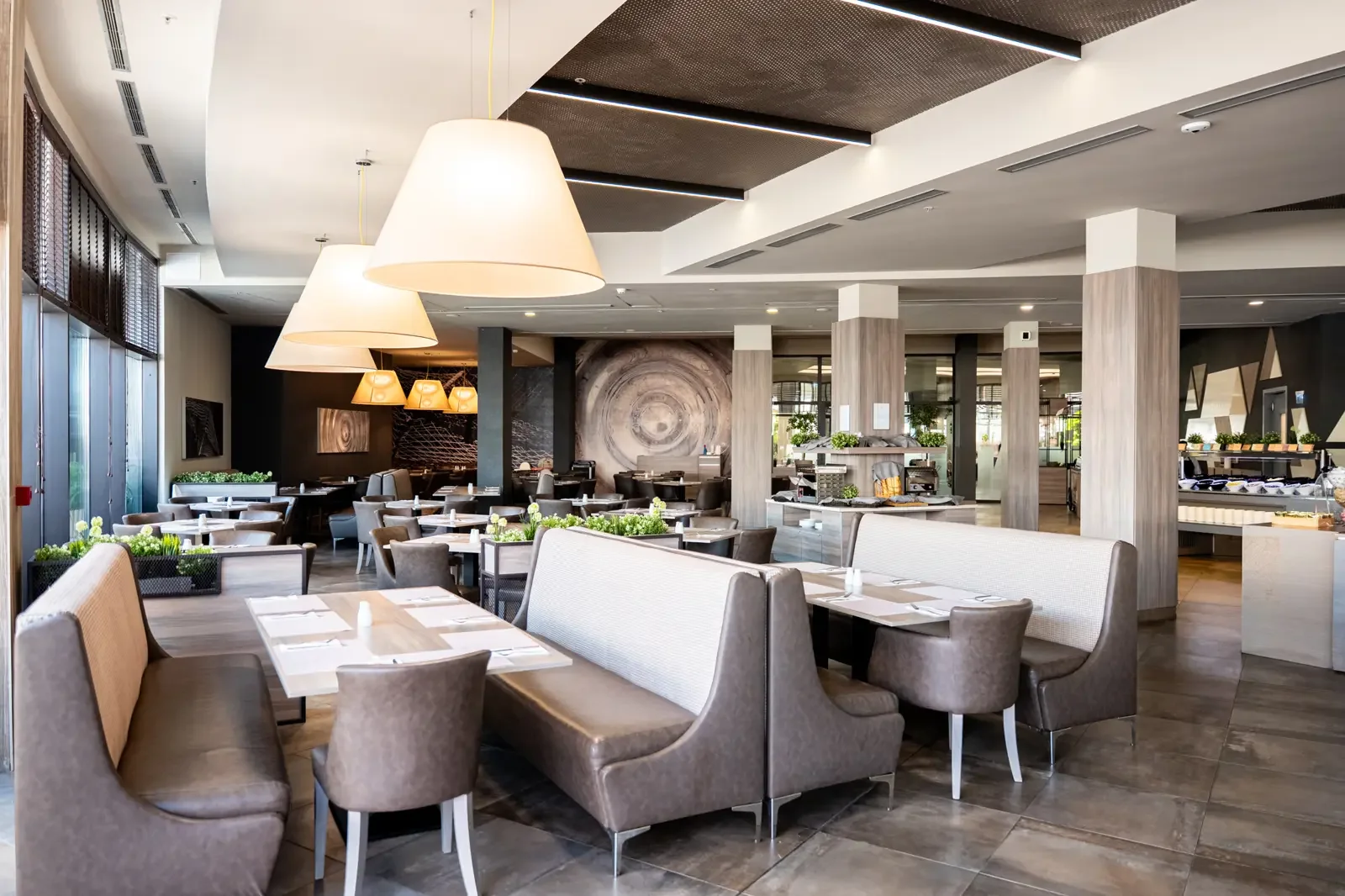 Interieur van een modern restaurant met diverse zitplaatsen, grote hangende lampen, decoratieve muurkunst, en veel natuurlijke lichtinval.
