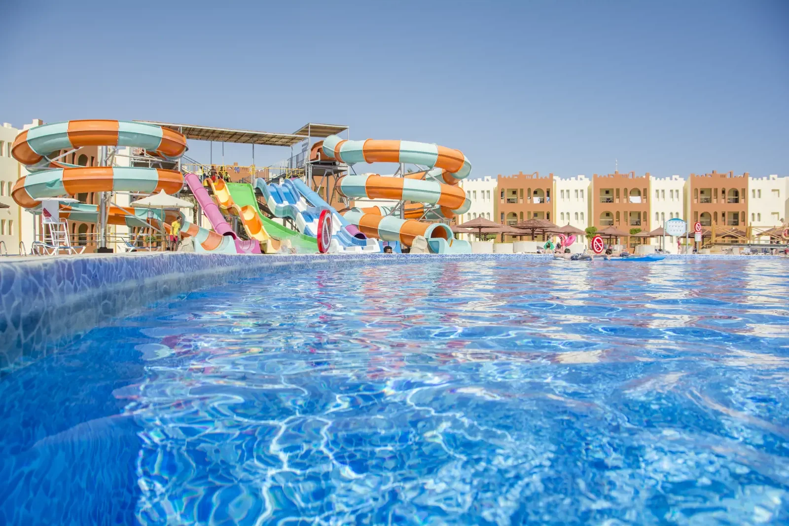 Een groot zwembadencomplex met meerdere waterslides in levendige kleuren, omgeven door ligbedden en een resort-achtig gebouw op de achtergrond, onder een heldere blauwe lucht.