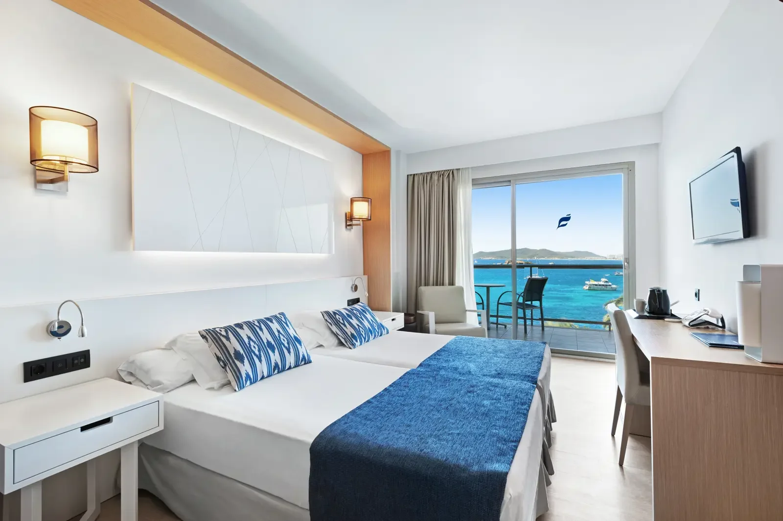 Hotelkamer met uitzicht op zee, tweepersoonsbed, zitje, balkon met tafel en stoel, televisie aan de muur, bureau met koffiemachine, waterkoker en landkaart, moderne inrichting, witte muren en houten accenten.