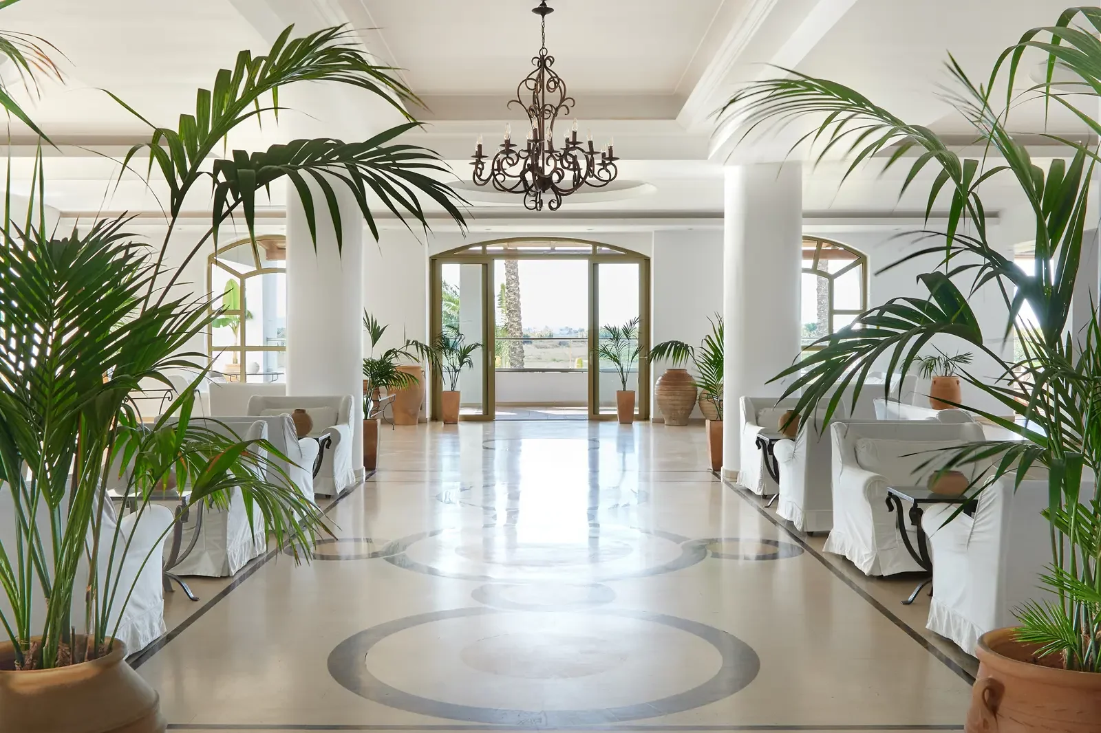 Luxe lobby met witte banken, planten in terracotta potten, grote ramen, marmeren vloer met patroon, kroonluchter aan het plafond.