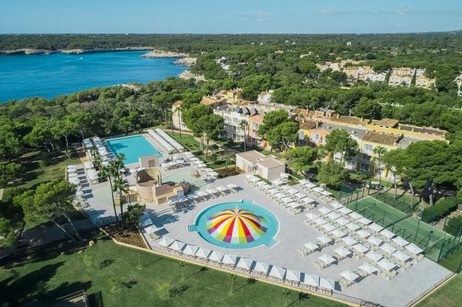 Een afbeelding van een zwembadcomplex met een kleurrijk waterballonvormig bad, omgeven door ligstoelen, palmbomen en een groene omgeving bij een resort, met een meer en bomen in de achtergrond.