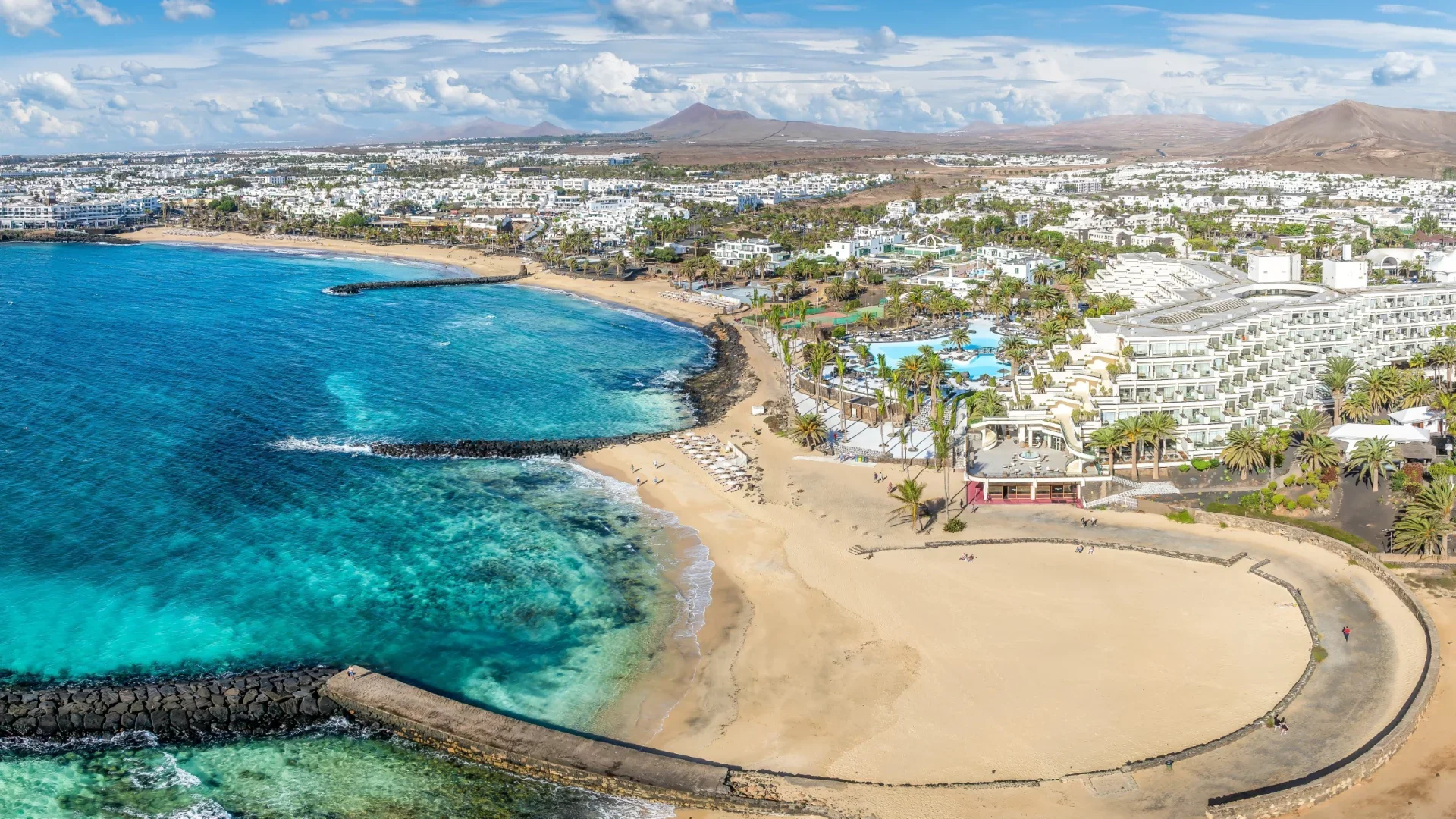 Een kustlijn met een zandstrand, blauwe oceaan en een modern resort met zwembaden en palmbomen, omgeven door een stedelijk gebied en bergen op de achtergrond.