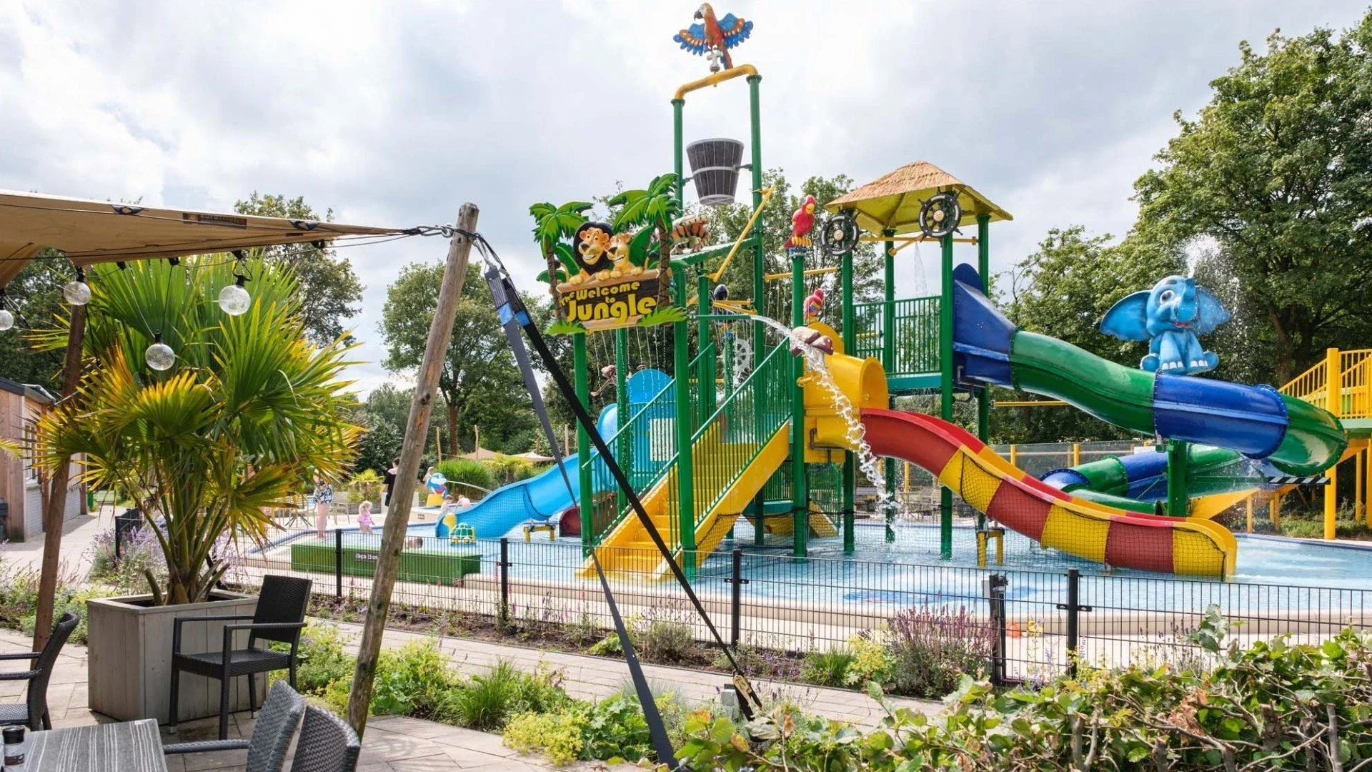 Kinderspeelplaats met waterattracties, glijbanen, en een waterpark, omgeven door groen en zitplaatsen.