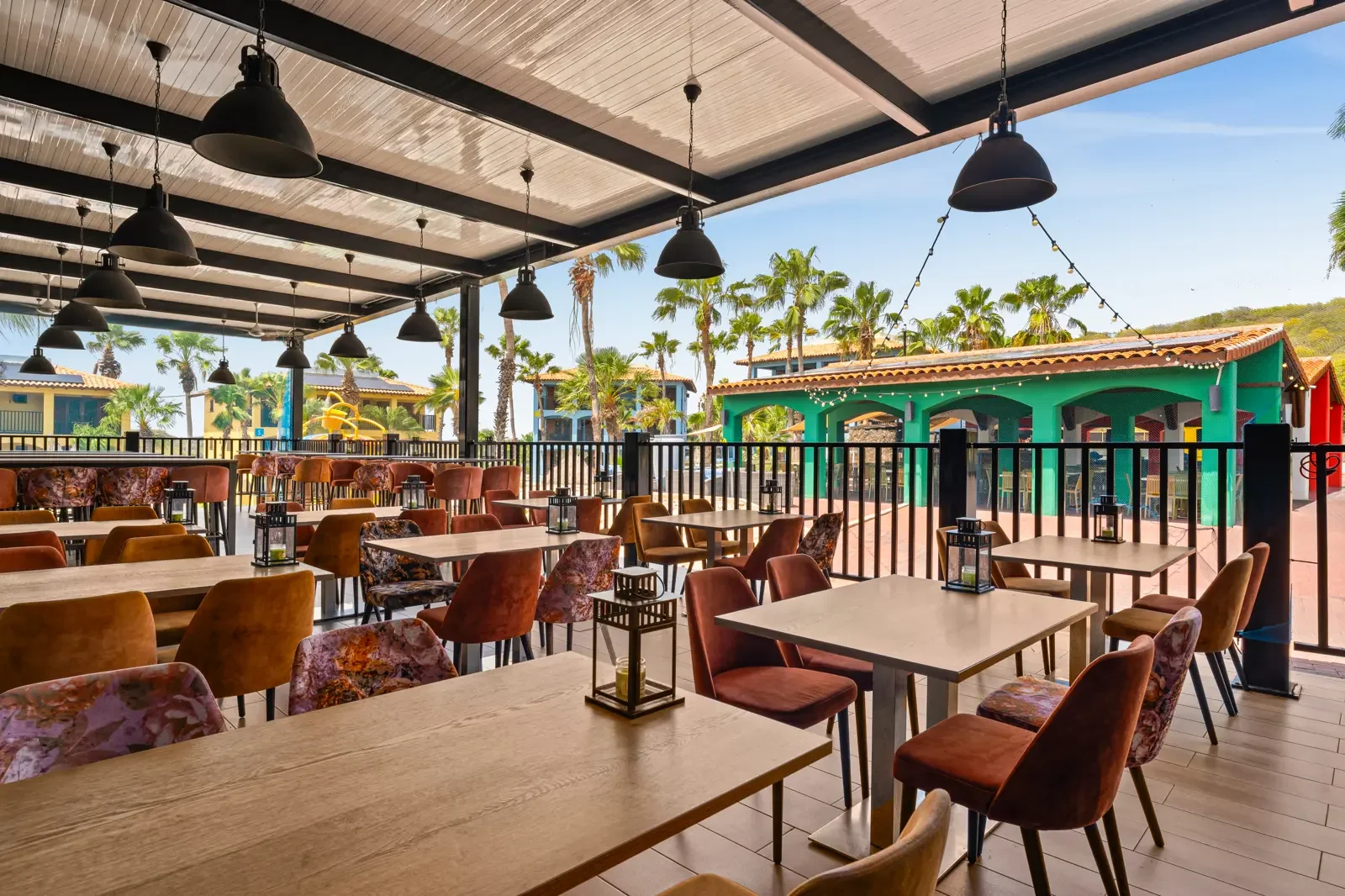 Binnenzicht van een restaurant met houten tafels en kleurrijke stoelen. Buiten is een speelplaats met veel palmbomen en speeltuig zichtbaar.
