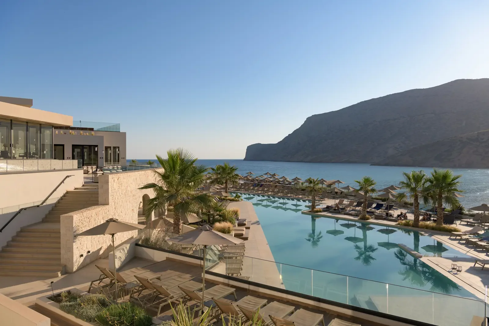 Luxe resort met infinitypool, ligstoelen, parasols, palm bomen en uitzicht op de zee en een rotsformatie op de achtergrond.