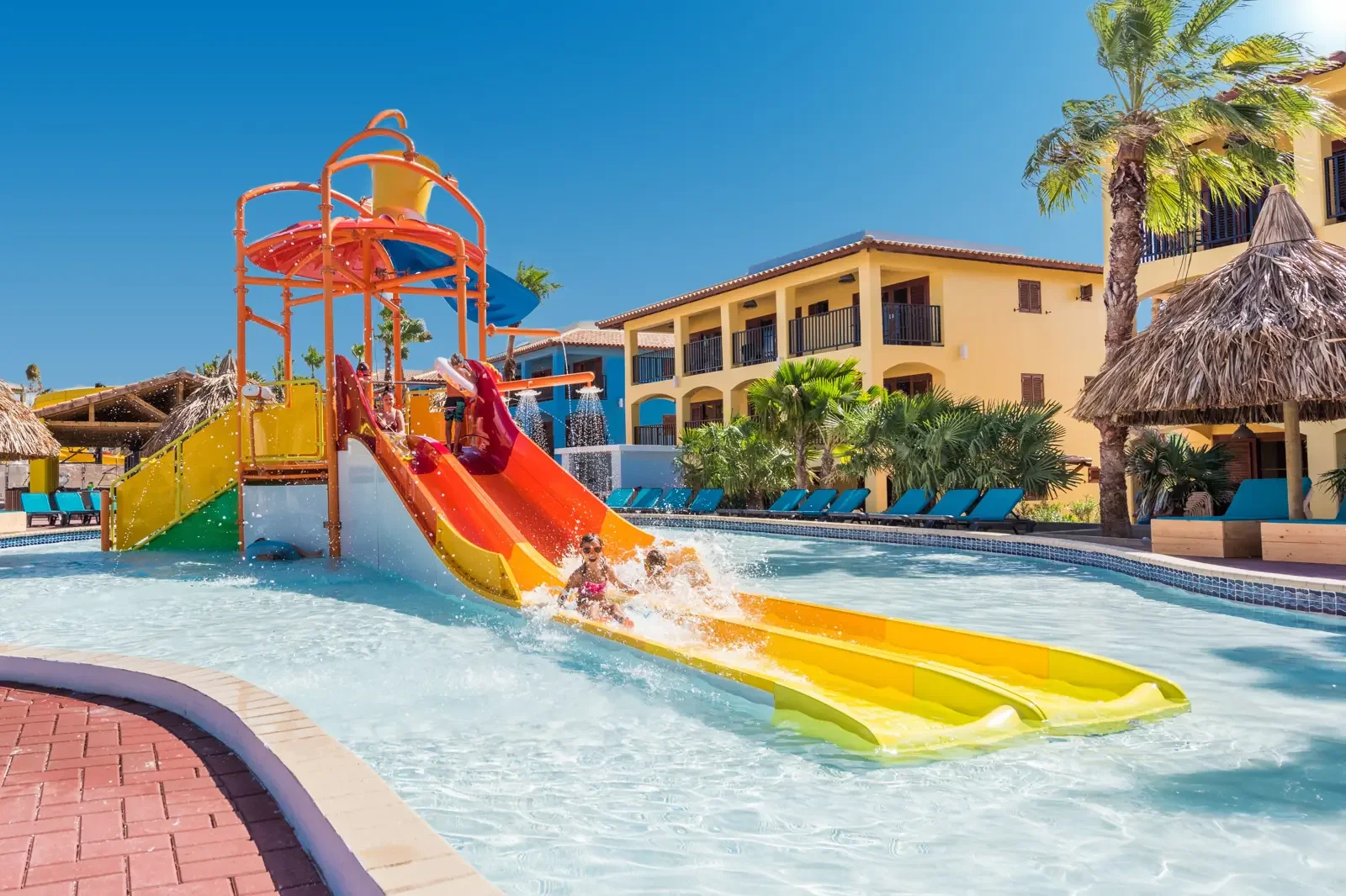 Kinder dieglijden in een waterpark met kleurrijke glijbanen, tropische palmbomen en een hotel met gele muren op de achtergrond onder een blauwe hemel.