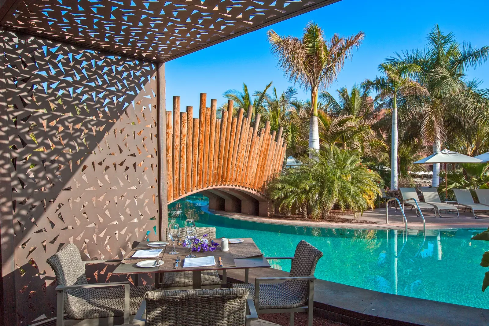 Een zwembad met ligbedden, parasols en veel palm bomen in een tropische setting. Er is een overdekte eethoek met een tafel en stoelen in de voorgrond. De omgeving is zonnig en blauw.