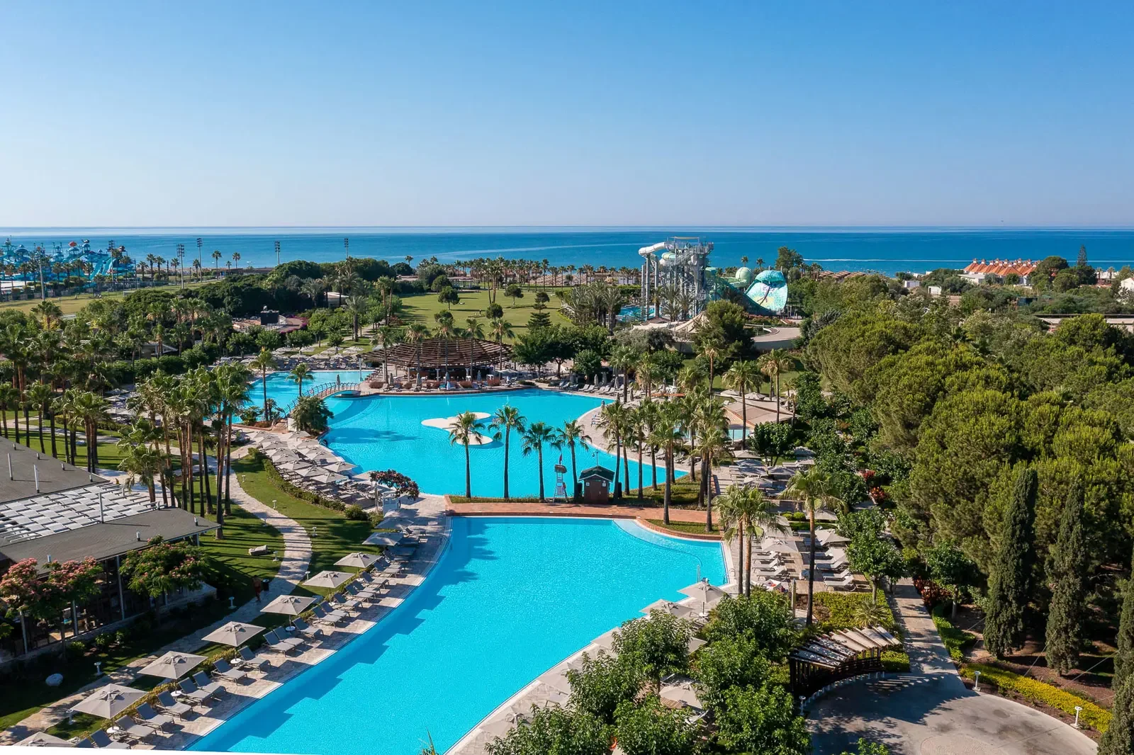 Luchtfoto van een waterpark met zwembaden, ligstoelen en palmbomen, met de oceaan op de achtergrond.