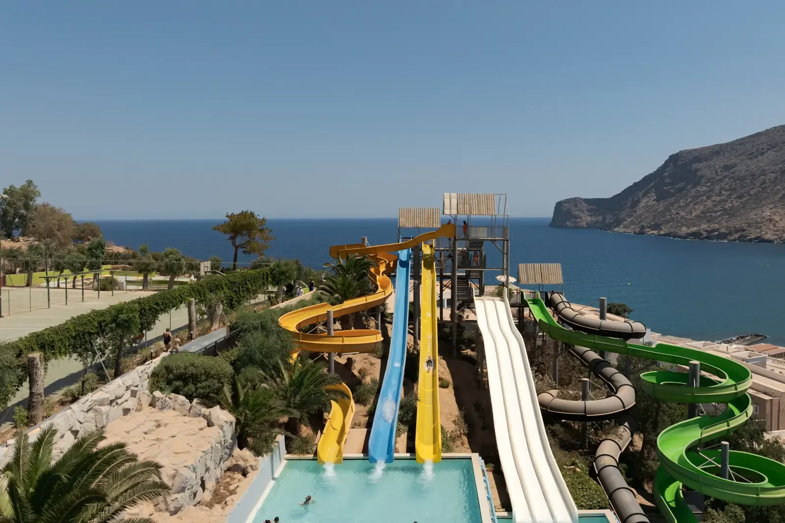 Foto van een waterpark met meerdere waterglijbanen, waaronder gele, blauwe, witte, groene en grijze, met een uitzicht op de zee en bergen op de achtergrond.
