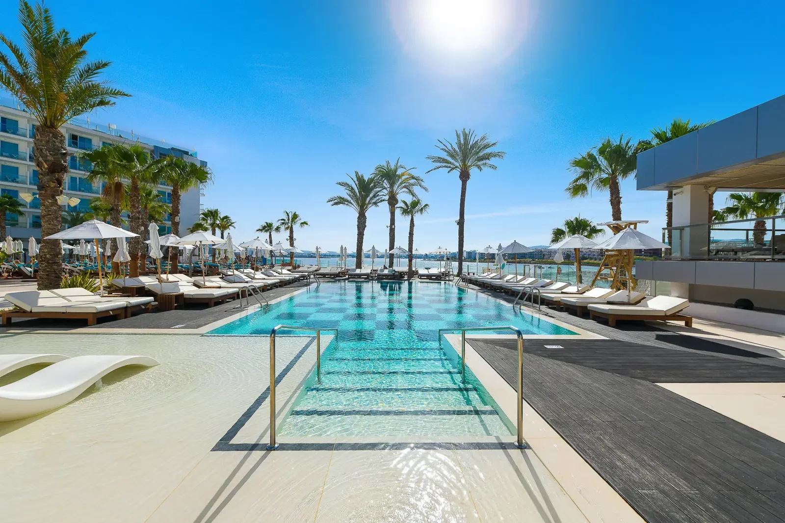 Luxe zwembad bij een hotel met ligstoelen en parasols, palmbomen en uitzicht op de oceaan op een zonnige dag.