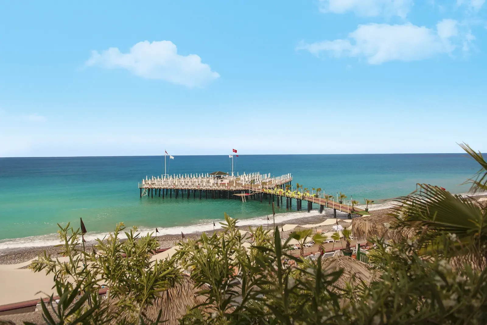 Een pier die uitsteekt in de zee met blauwe lucht en wat wolken, omringd door een strand en palmbomen.