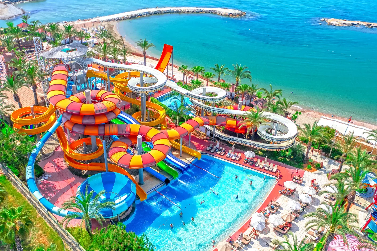 Een waterpark met meerdere kleurrijke glijbanen en een zwembad, gelegen aan de kust met een strand en de zee op de achtergrond.