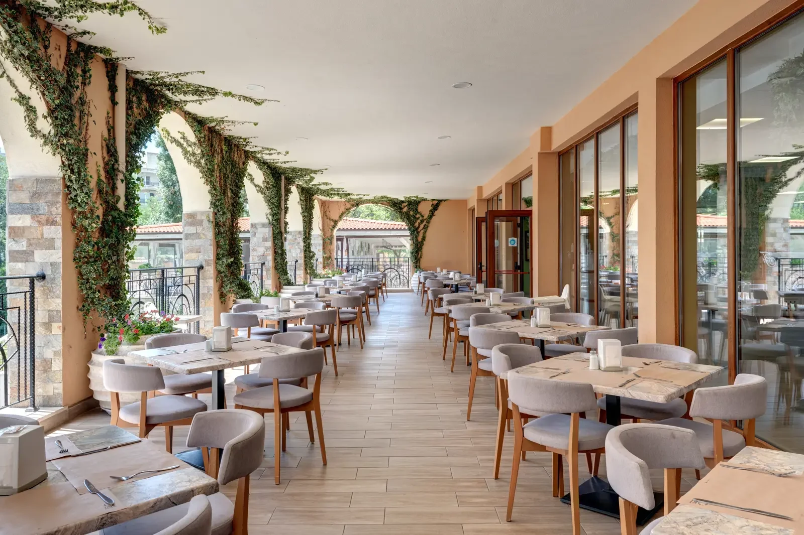 Een overdekt terras van een restaurant met rijen witte en beige stoelen, tafels met servetten en bestek, grote ramen, planten, en een uitzicht op buiten.