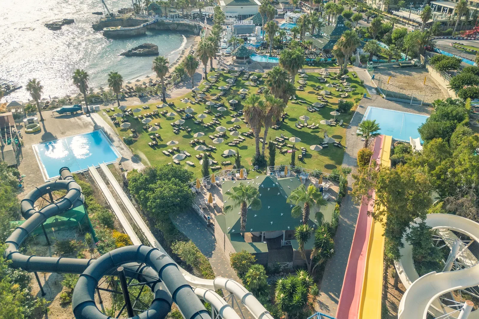 Luchtfoto van een waterpark met glijbanen, zwembaden, ligbedden, parasols, palmbomen en een strand met een rots in de zee.