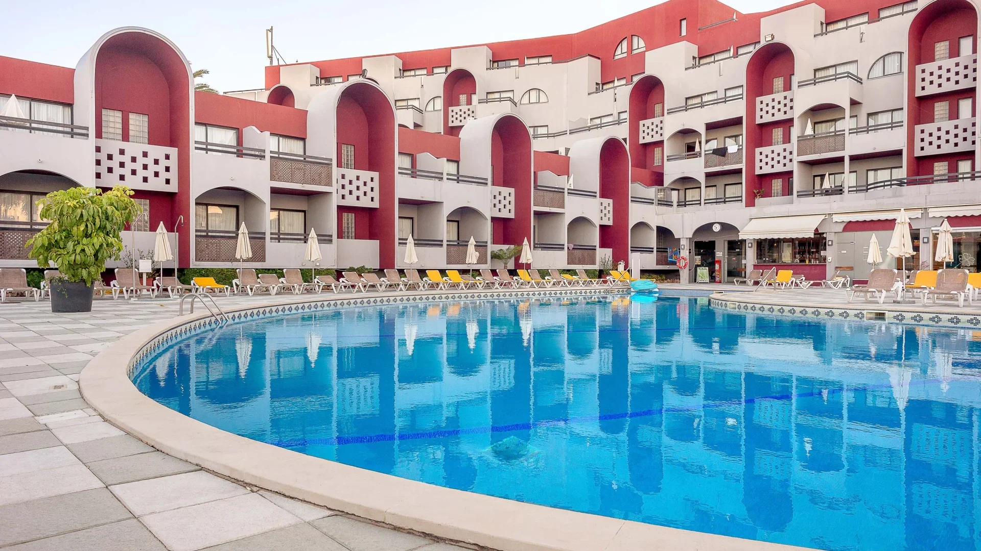 Een zwembad met ligstoelen en parasols voor een wit en rood hotelgebouw met balkons en ronde ramen.