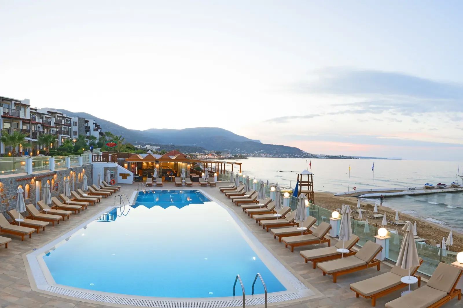 Zwembad met ligbedden en parasols bij een strandhotel met uitzicht op de zee, zonsondergang, boten en bergen op de achtergrond.